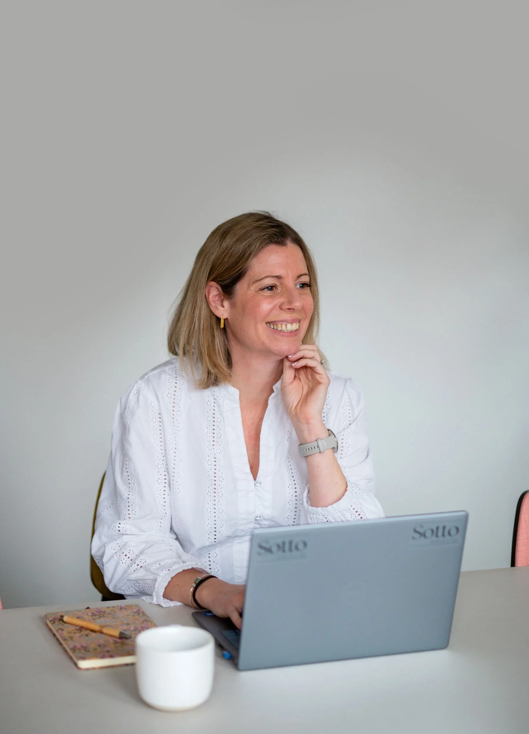A woman with shoulder-length blonde hair, wearing a white blouse, smiling while working on a laptop at a desk. There is a white mug and a floral notebook with a pen on the desk.