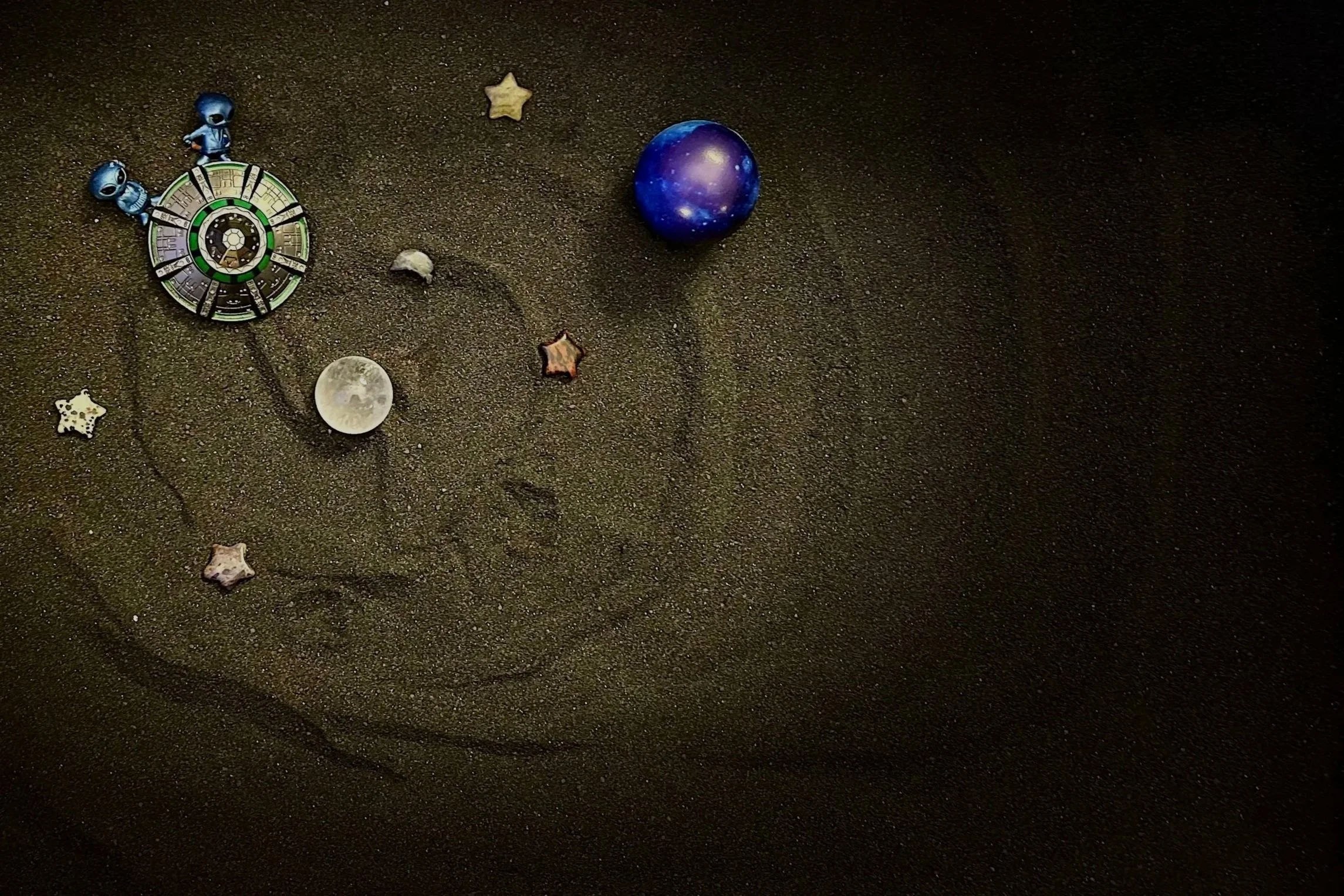 An image of a sandtray scene made in black sand depicting two aliens riding a spaceship among stars and planets.