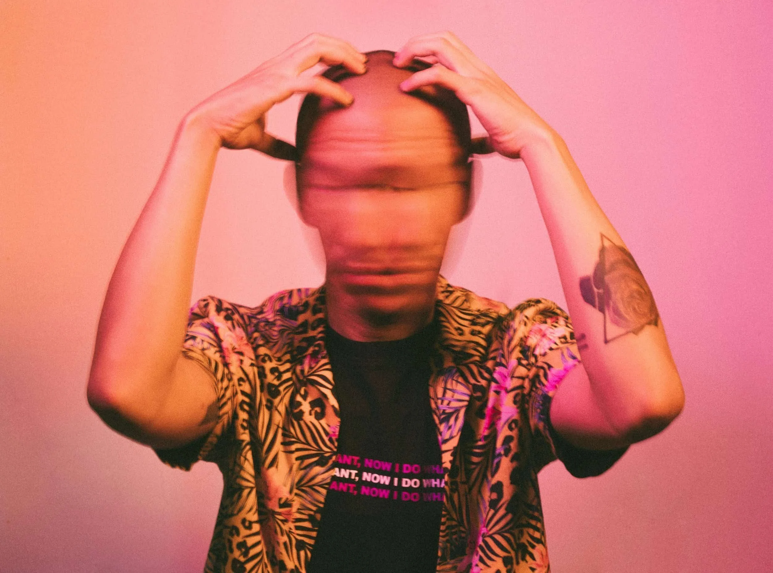 A person sitting in front of a pink background. They have both of their hands on their head. Their head is blurred, suggesting they are shaking it back and forth.