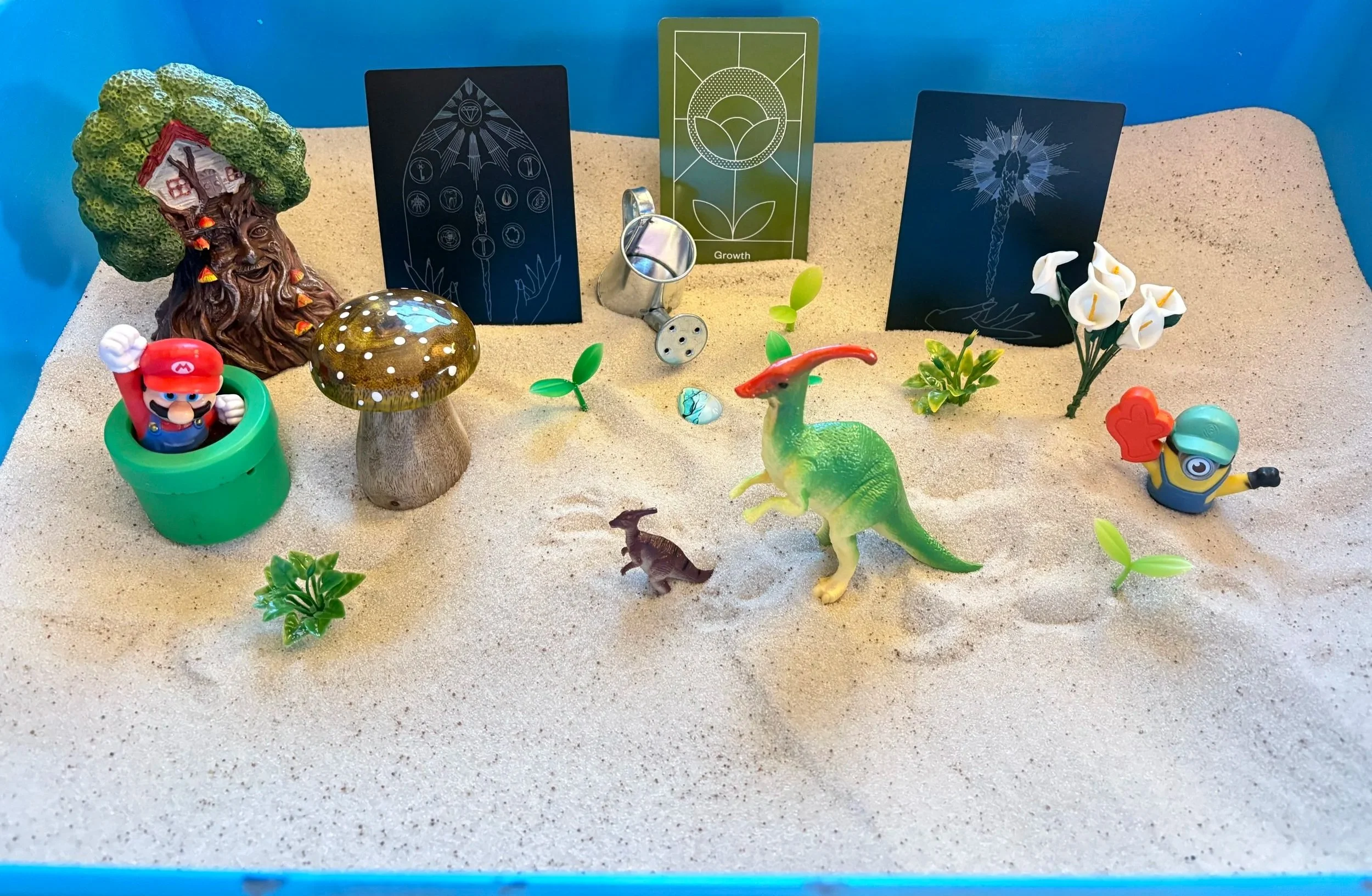 A sandtray scene using a light blue sandtray with white sand. The scene shows elements of nature, tarot and oracle cards with the theme of growth, miniatures of Mario and a minion looking triumphant, and two similar dinosaurs of different sizes.