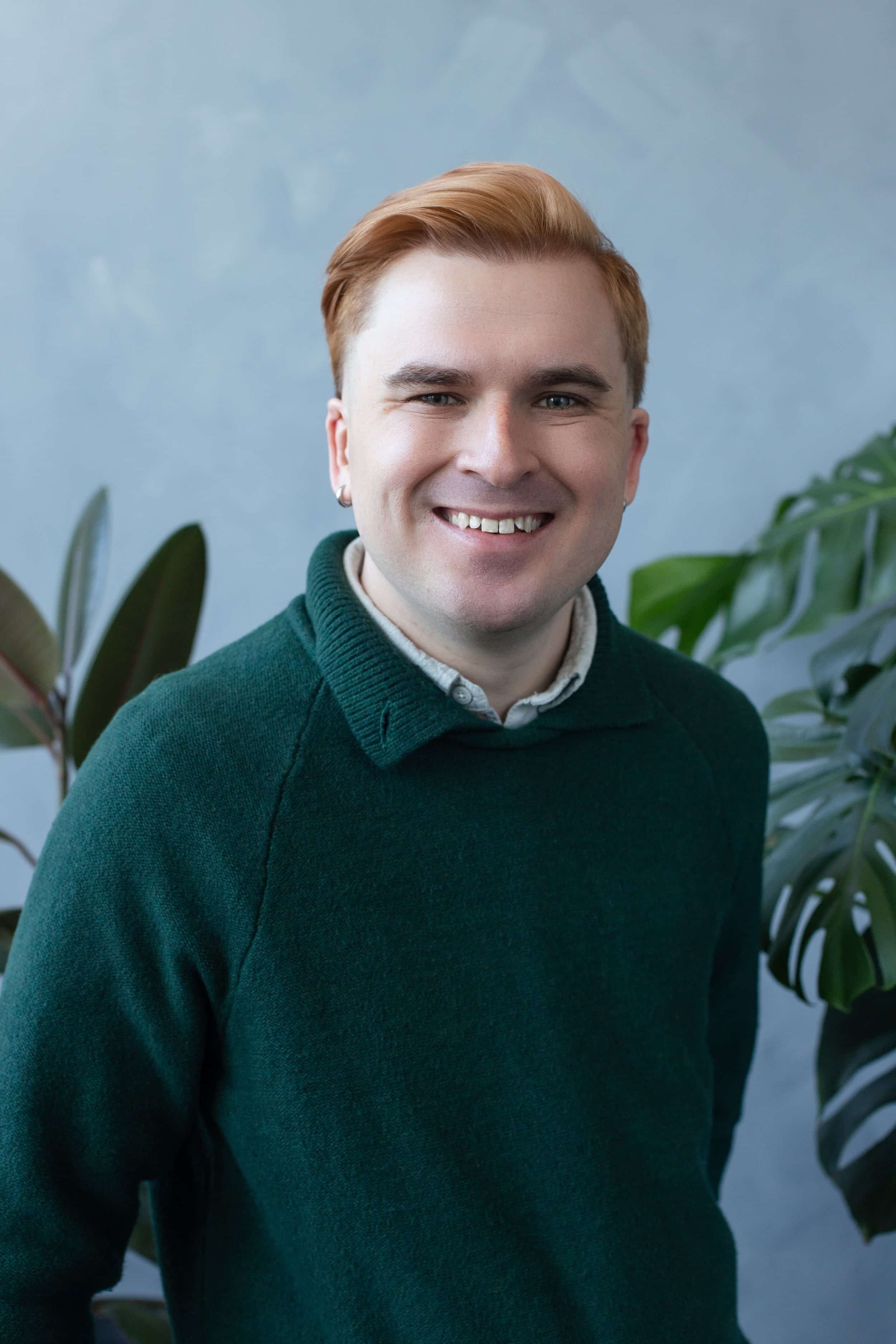 Headshot of therapist Elijah Eiler, LCSW. Elijah is looking into the camera and smiling. Elijah is wearing a green sweater and standing in front of a blue background with plants behind him.