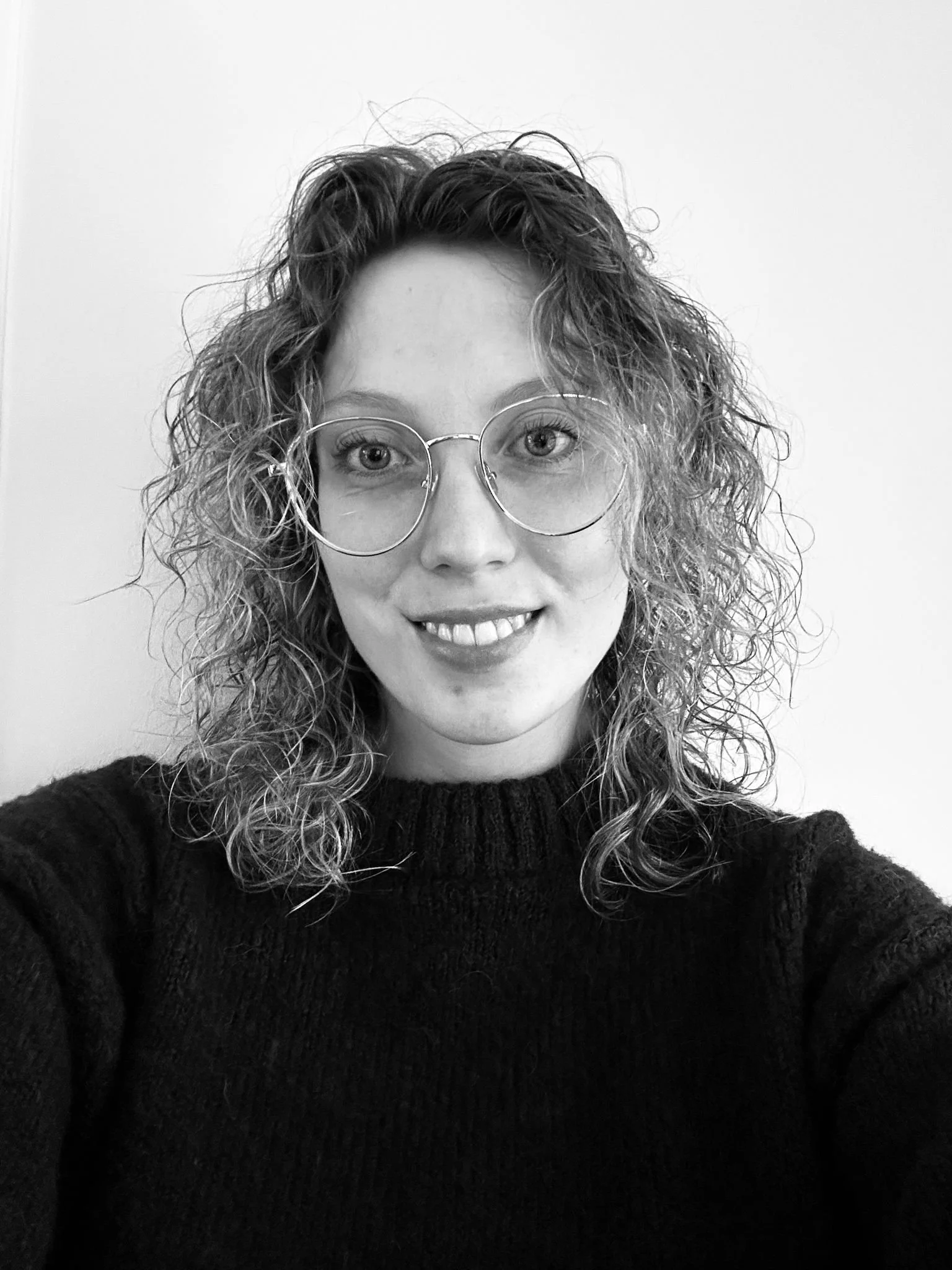 A woman with curly blonde hair and glasses smiling at the camera, seated on a white chair with a light-colored curtain behind her.