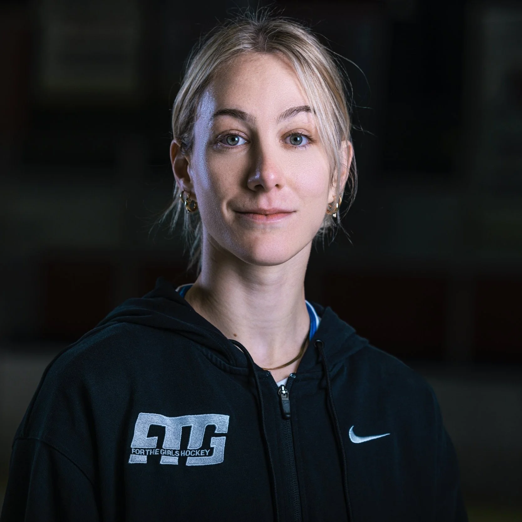 A portrait of a young woman with blonde hair, blue eyes, and gold hoop earrings, wearing a black hoodie, looking directly at the camera with a slight smile.