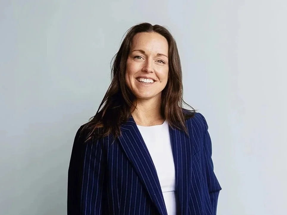 A woman with brown hair, wearing a navy pinstripe blazer and white top, smiling in front of a light background.
