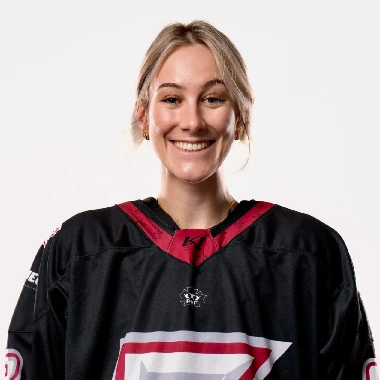 A portrait of a young woman with blonde hair, blue eyes, and gold hoop earrings, wearing a black hoodie, looking directly at the camera with a slight smile.