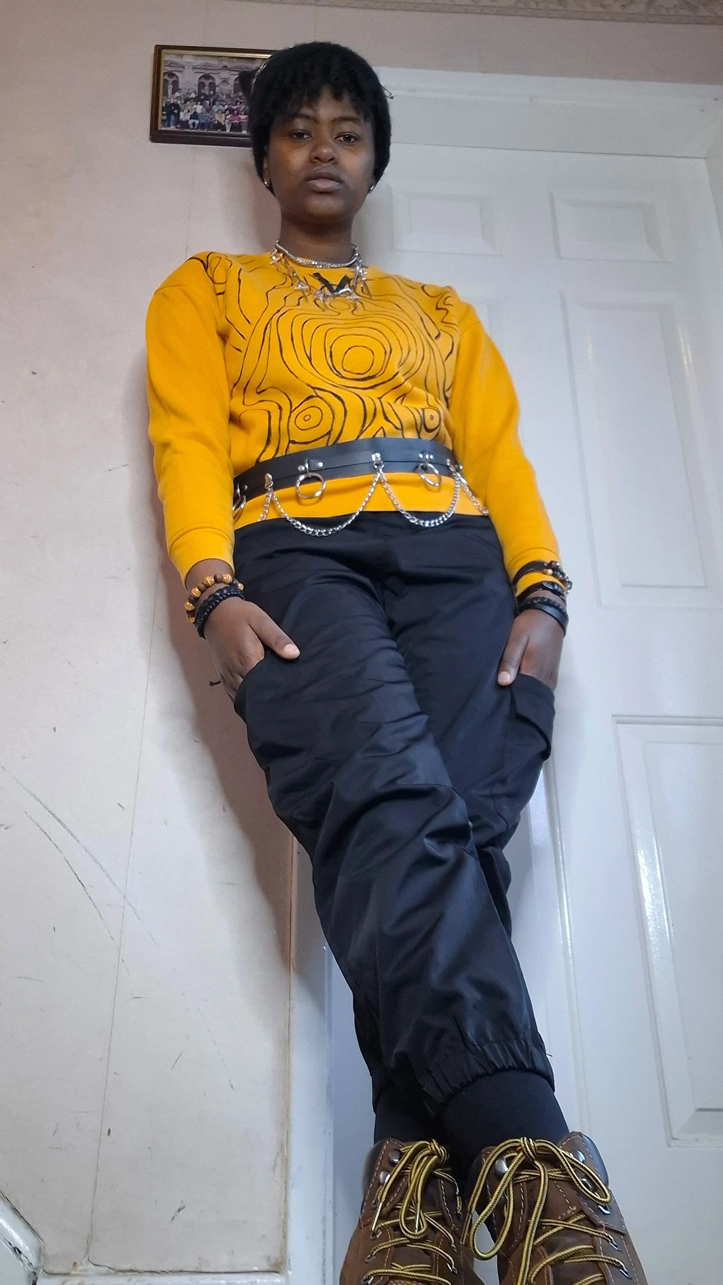 a girl wearing a  black beanie cap, a yellow  pullover with black design, a black and silver belt, black cargo pants, black and yellow bracelets, and silver necklaces