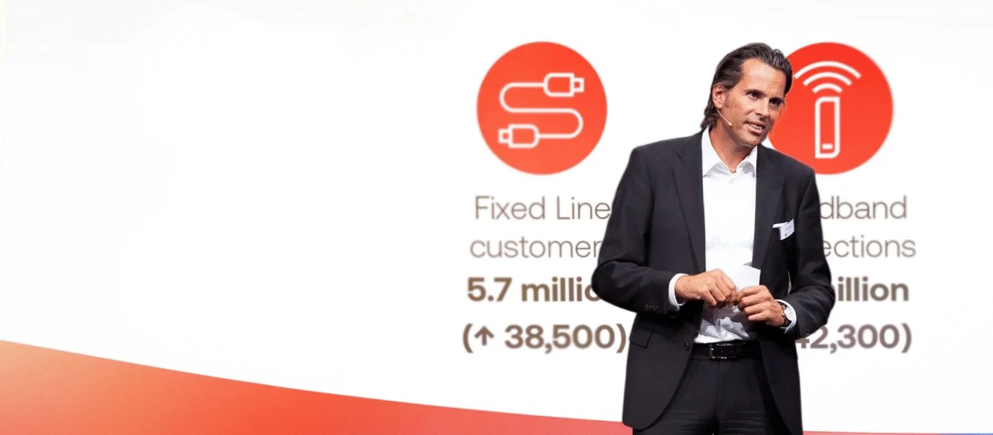 A man in a dark suit giving a presentation in front of a slide with icons of cables and a mobile device, and text about fixed line and broadband customer growth.