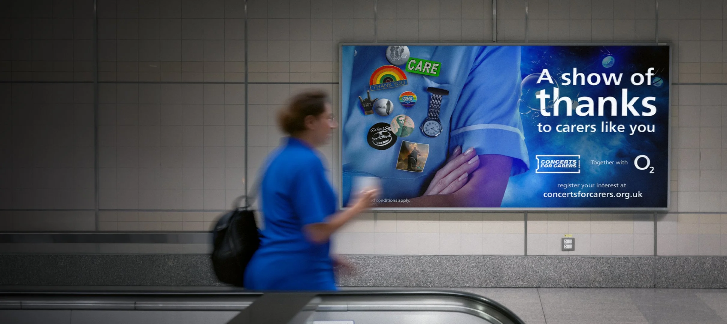 A person in blue clothing walking past a billboard in an indoor setting, with the billboard displaying messages of thanks to carers and various badges and pins.