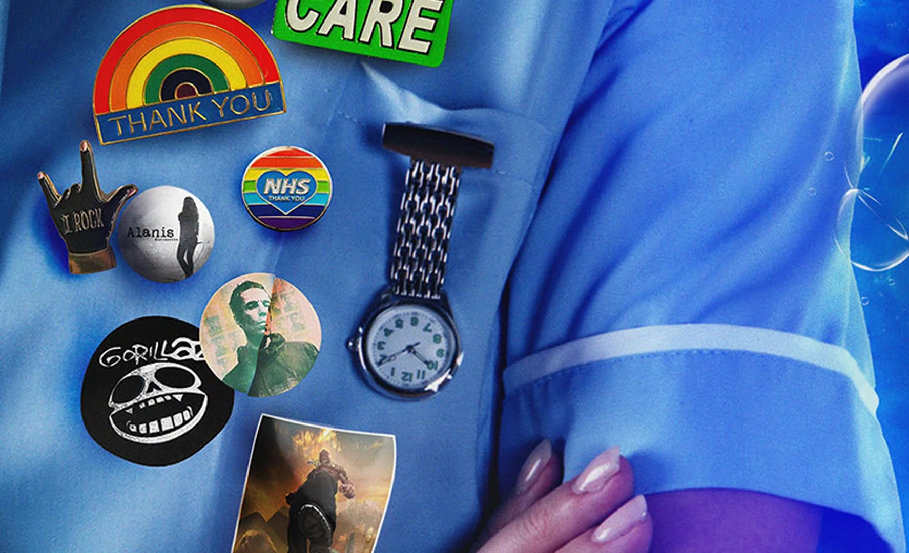 Close-up of a person's torso wearing a blue shirt with various buttons, patches, and stickers. Notable items include a rainbow sticker with 'Thank You', a rainbow NHS badge, a black patch with a skull, a round sticker with a portrait of a woman, and an analog watch with a black and white striped strap.