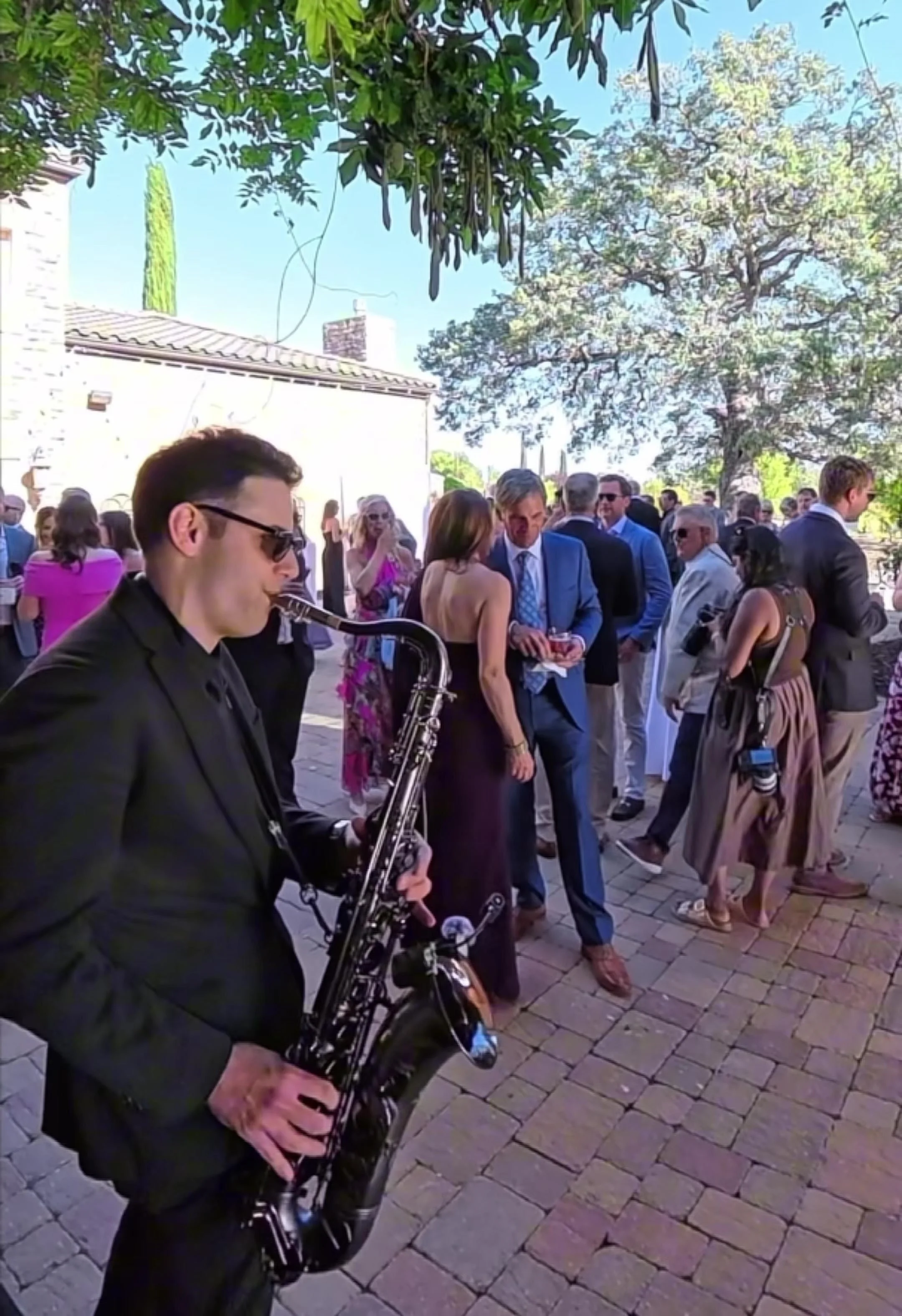 Ant Smith playing saxophone while guests mingle during an outdoor wedding cocktail hour.