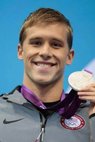 Young male athlete smiling and holding a silver medal with a purple ribbon, wearing a gray jacket with a USA Olympic team logo.