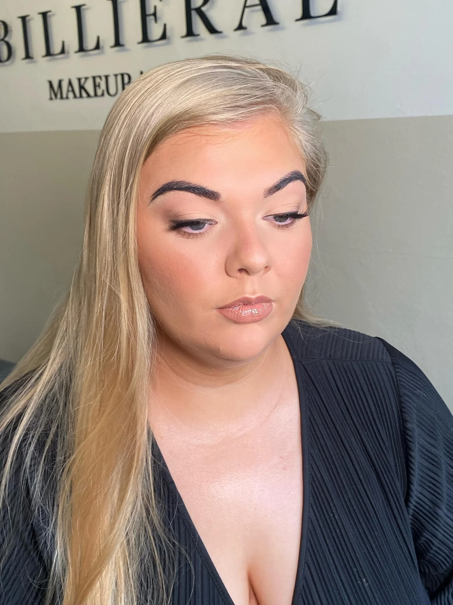 Close-up of a woman with long blonde hair and makeup, looking downward. Background has part of a sign that reads 'BILLIERE MAKEUP'.