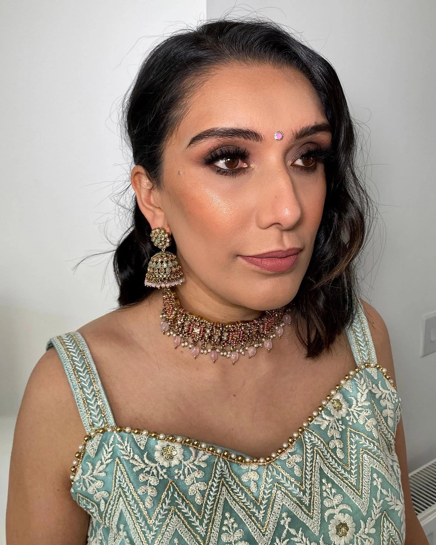 A woman with dark hair styled in loose waves, wearing traditional Indian jewelry including chandelier earrings, a necklace, and a small bindi on her forehead, dressed in a light blue embroidered outfit.