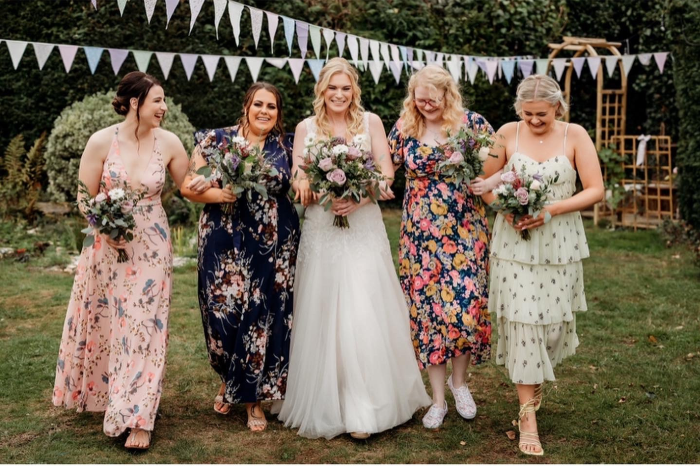 Six women, including a bride in a white wedding dress, walking arm-in-arm outdoors at a wedding reception or celebration. They are smiling and holding bouquets, with triangular bunting decorations overhead and greenery in the background.