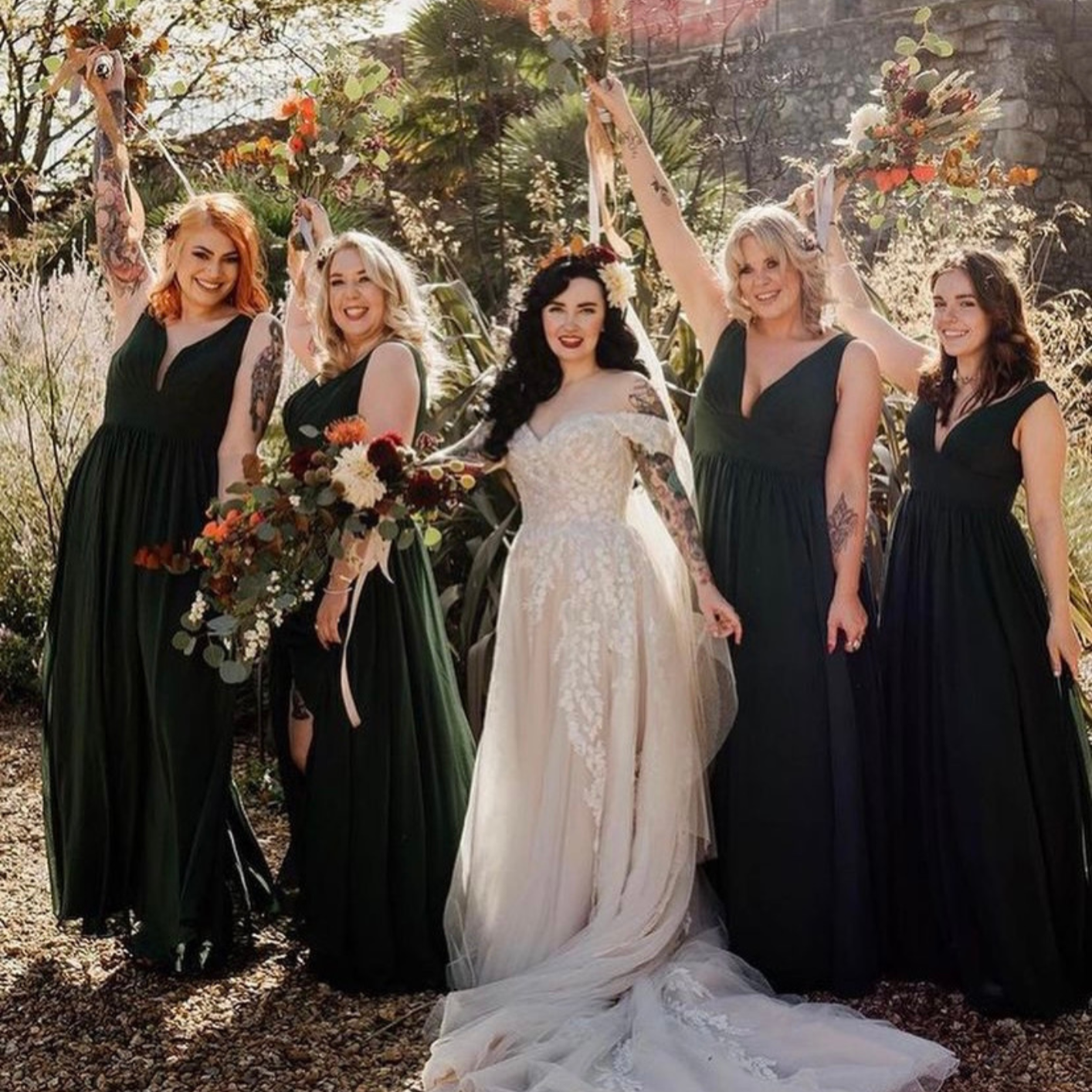 A bride in a white wedding dress holding a bouquet of flowers, surrounded by five bridesmaids in black dresses, outdoors with trees and plants in the background.
