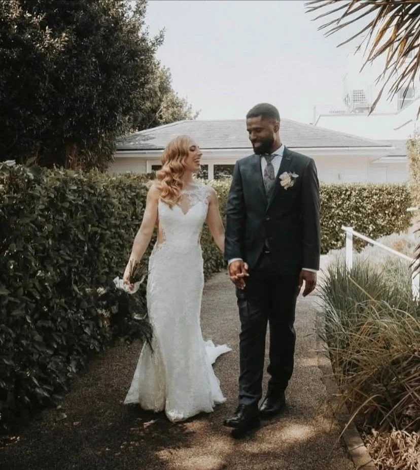 Bride and groom walking hand in hand outdoors, holding hands, with the bride in a white wedding gown and the groom in a dark suit, surrounded by bushes and a house in the background.