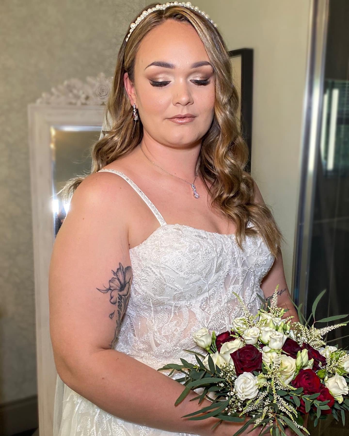 Woman in wedding dress holding a bouquet of white and red roses, with a tattoo on her left arm and wearing jewelry, standing indoors.