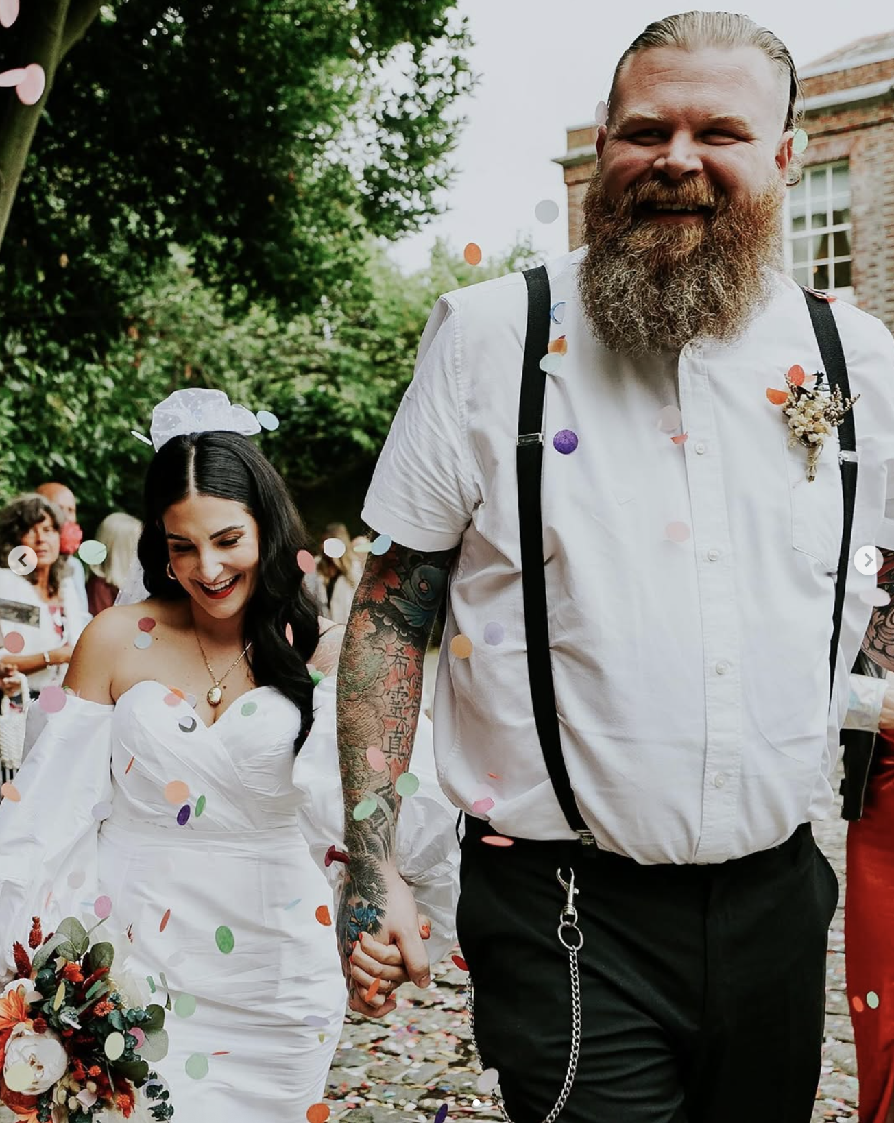 A bride and groom holding hands at their wedding, surrounded by colorful confetti and guests, outdoors with greenery and a brick building in the background.
