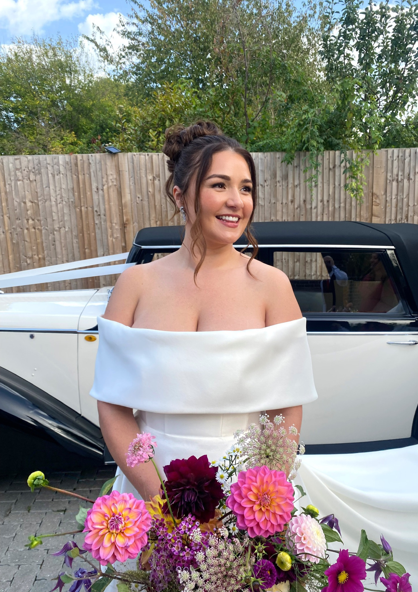 A smiling bride in a white off-shoulder wedding dress holding a colorful bouquet of flowers, standing outdoors in front of a vintage car and a wooden fence.