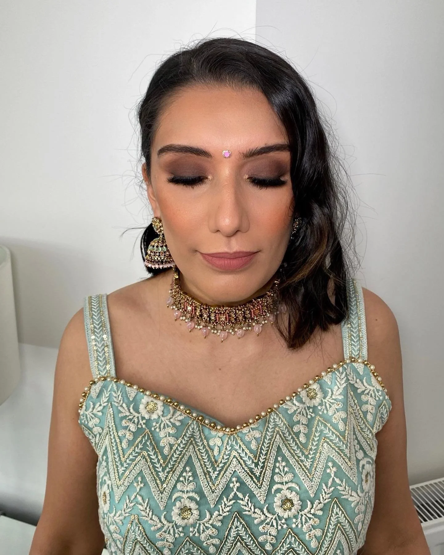 A woman with dark, wavy hair and closed eyes, dressed in traditional Indian attire with jewelry and makeup, standing against a plain white background.