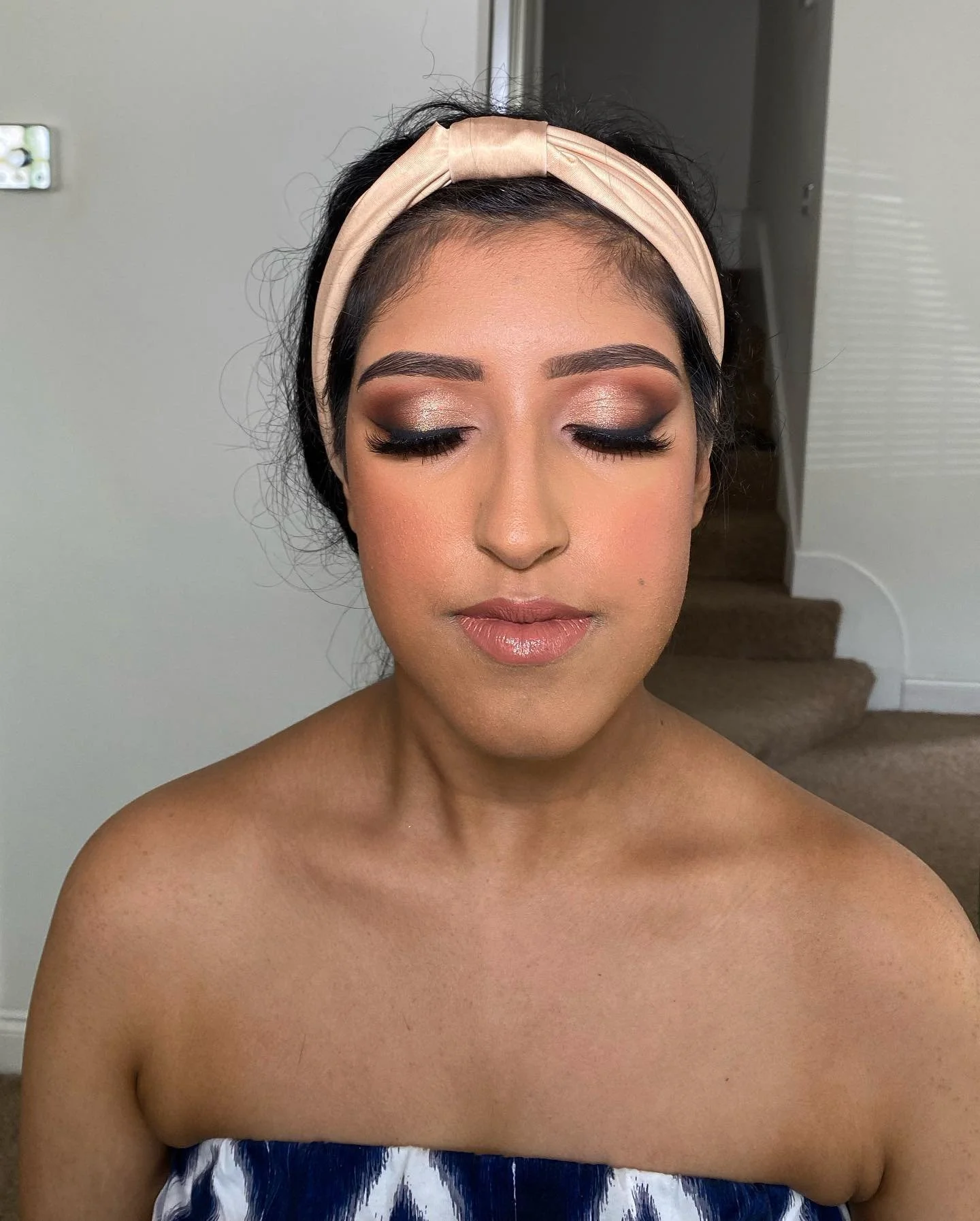 Woman with makeup and a light pink headband, closed eyes, standing indoors, with stairs in the background.