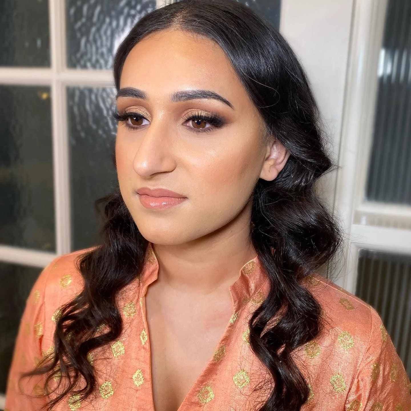 A woman with tan skin, long dark wavy hair, and makeup featuring gold eyeshadow, long eyelashes, and glossy lips, wearing a peach-colored traditional dress with gold embroidery, standing indoors in front of windows.