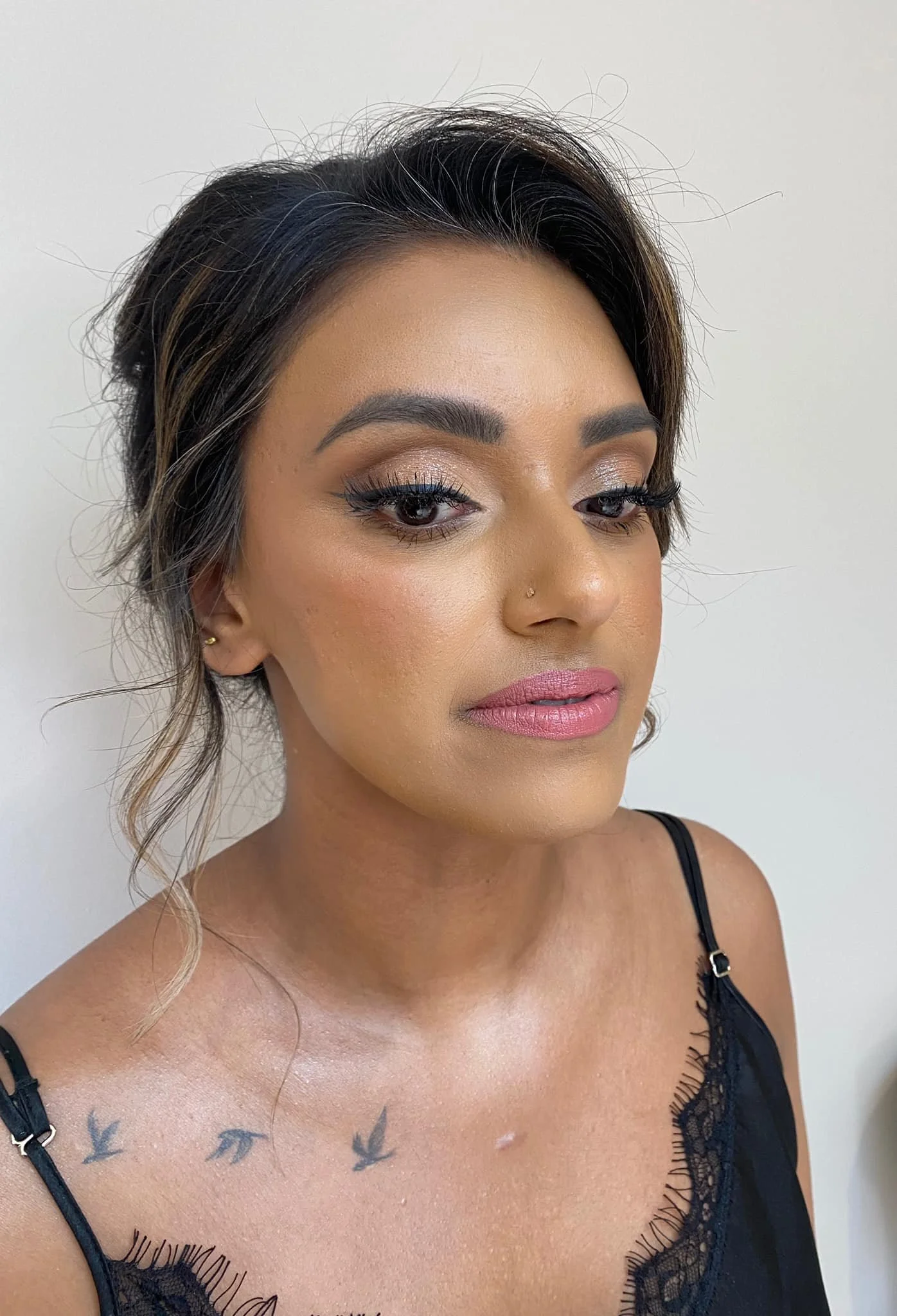 A woman with dark, styled hair, eye makeup, pink lipstick, and a nose piercing, wearing a black lace camisole, standing against a plain white wall.