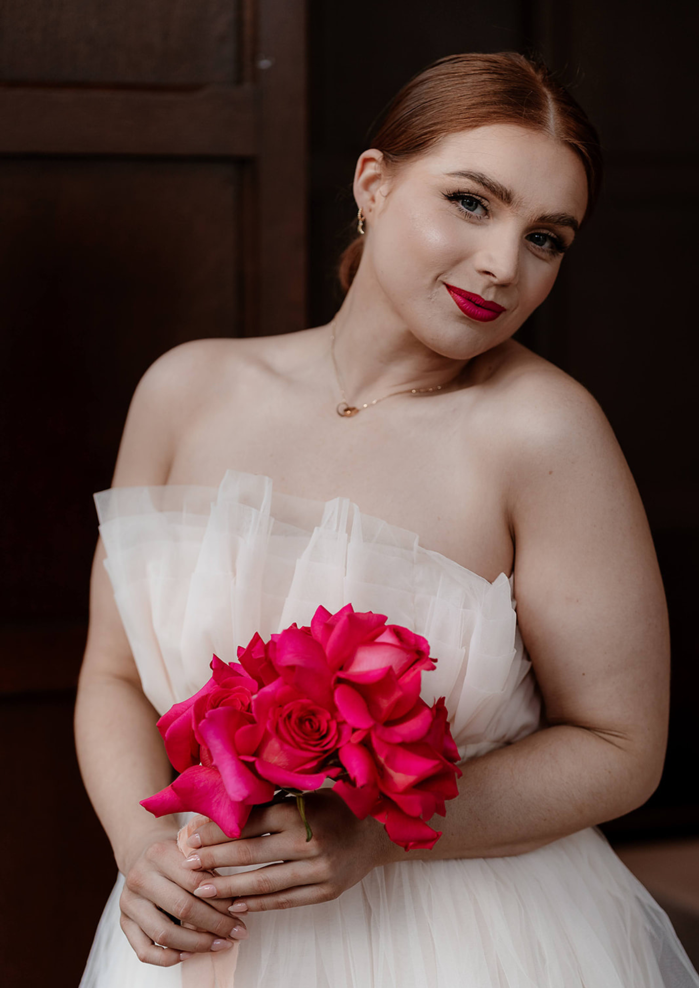 A woman with red hair and makeup is holding a bouquet of bright pink roses and wearing a strapless white dress, standing in front of a dark wooden background.