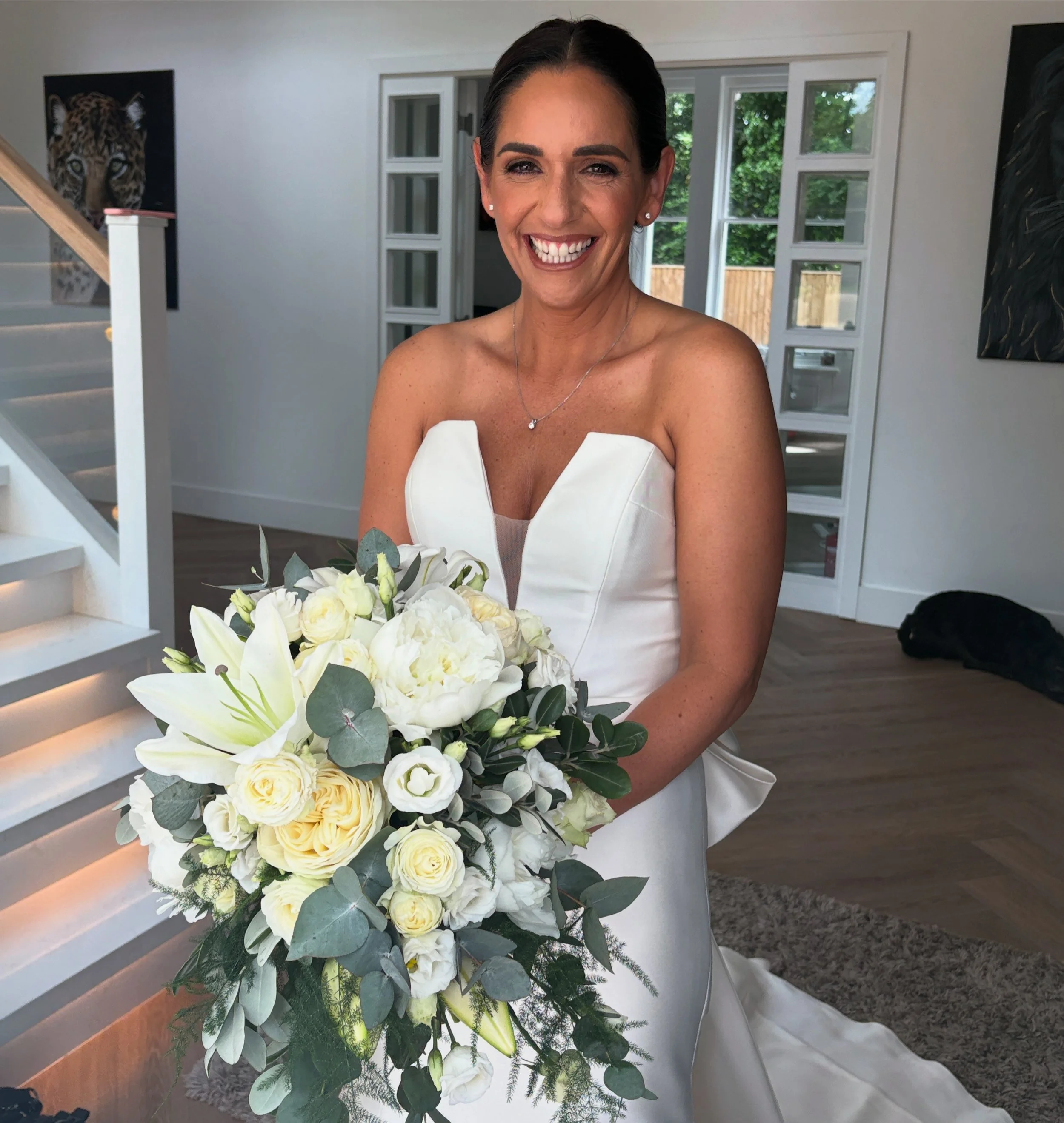 A woman in a white wedding dress holding a large bouquet of white flowers, smiling indoors with a staircase and artwork on the walls behind her.