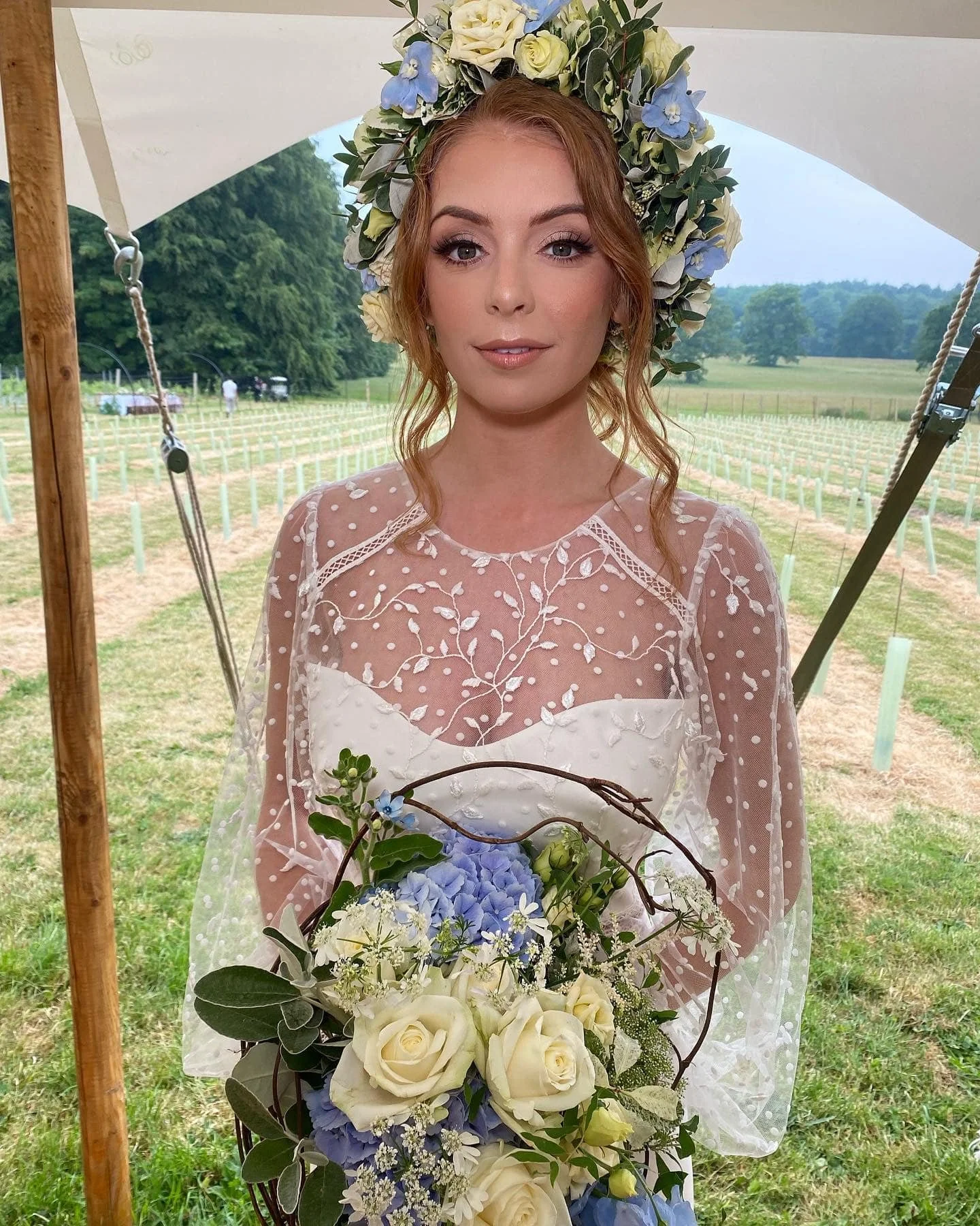 A woman wearing wedding attire with a floral crown and holding a bouquet of white roses, blue flowers, and greenery, standing under a tent in a grassy outdoor setting.