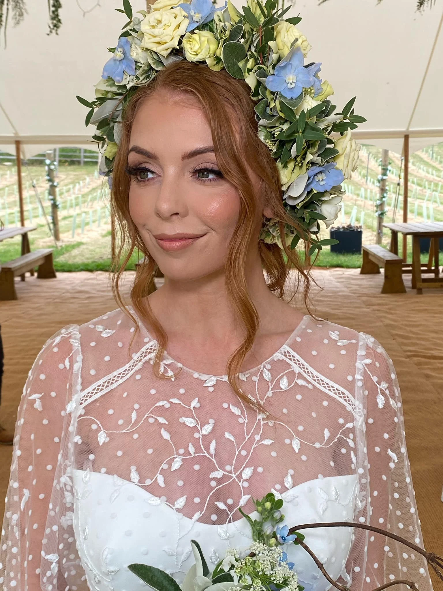 A woman with red hair wearing a floral crown of white, blue, and green flowers. She is dressed in a white lace dress with embroidered floral designs and is holding a small bouquet of white flowers and greenery. She is inside a tent with wooden benche