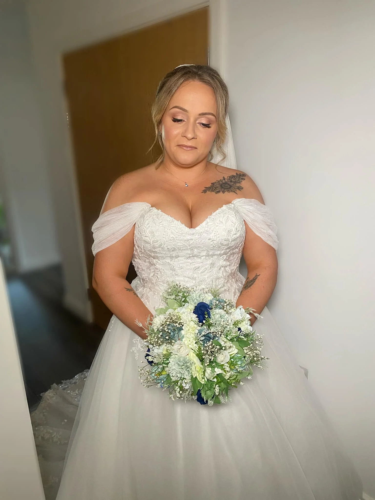 Bride in a white wedding dress holding a bouquet of white and blue flowers, standing indoors with a neutral expression.