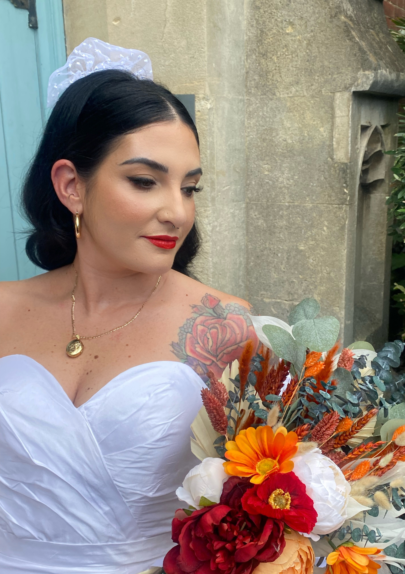 A woman with black hair, wearing red lipstick, gold hoop earrings, and a gold necklace, holding a colorful bouquet of flowers outdoors in front of a stone wall.