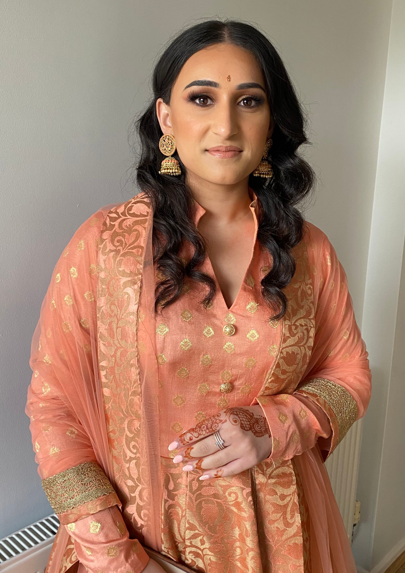 A woman dressed in traditional Indian attire, wearing a peach-colored embroidered suit, gold earrings, and a small bindi on her forehead, with henna designs on her hand.