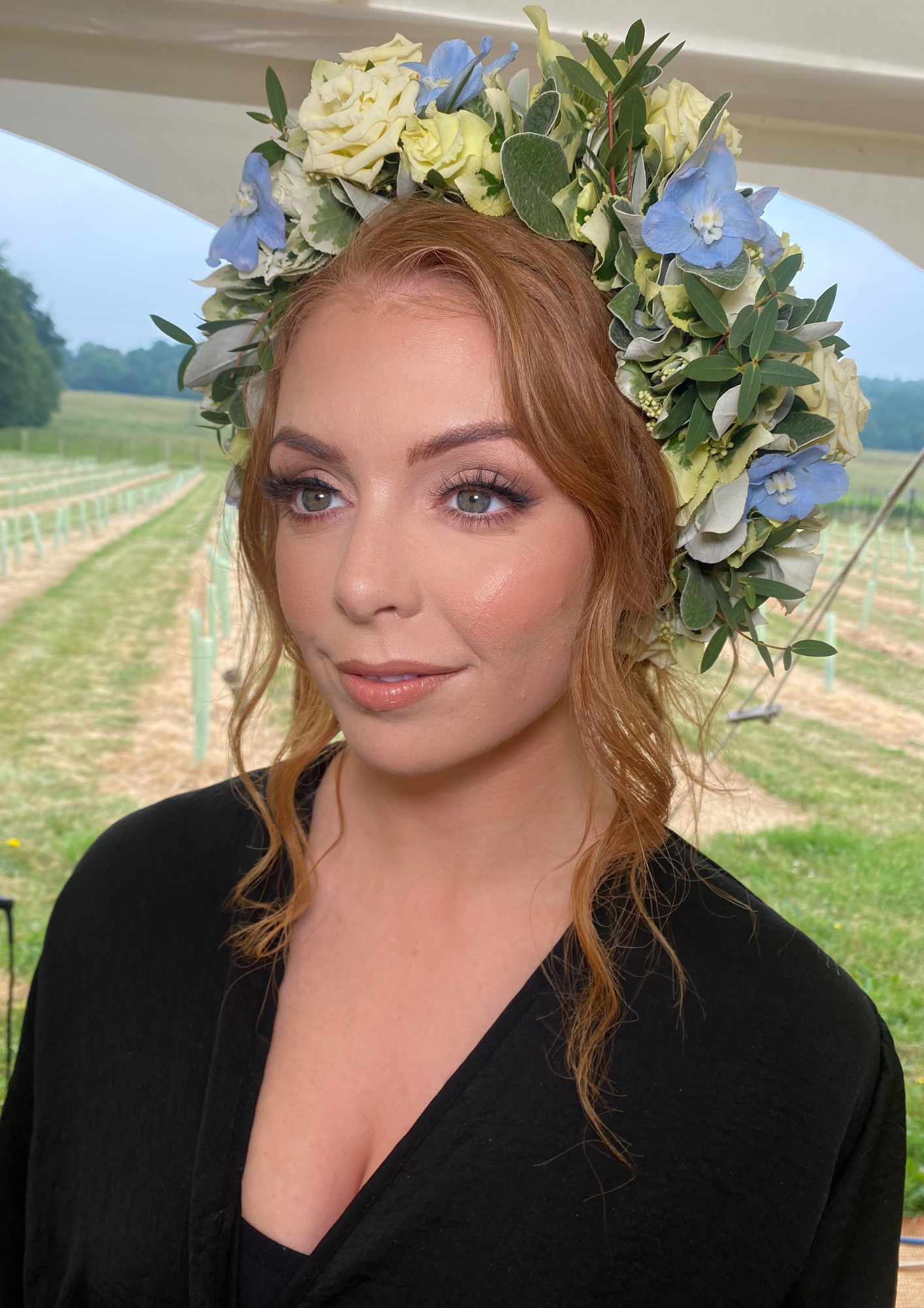A woman with red hair and makeup wearing a floral crown made of white, green, and blue flowers, standing outdoors in a field.
