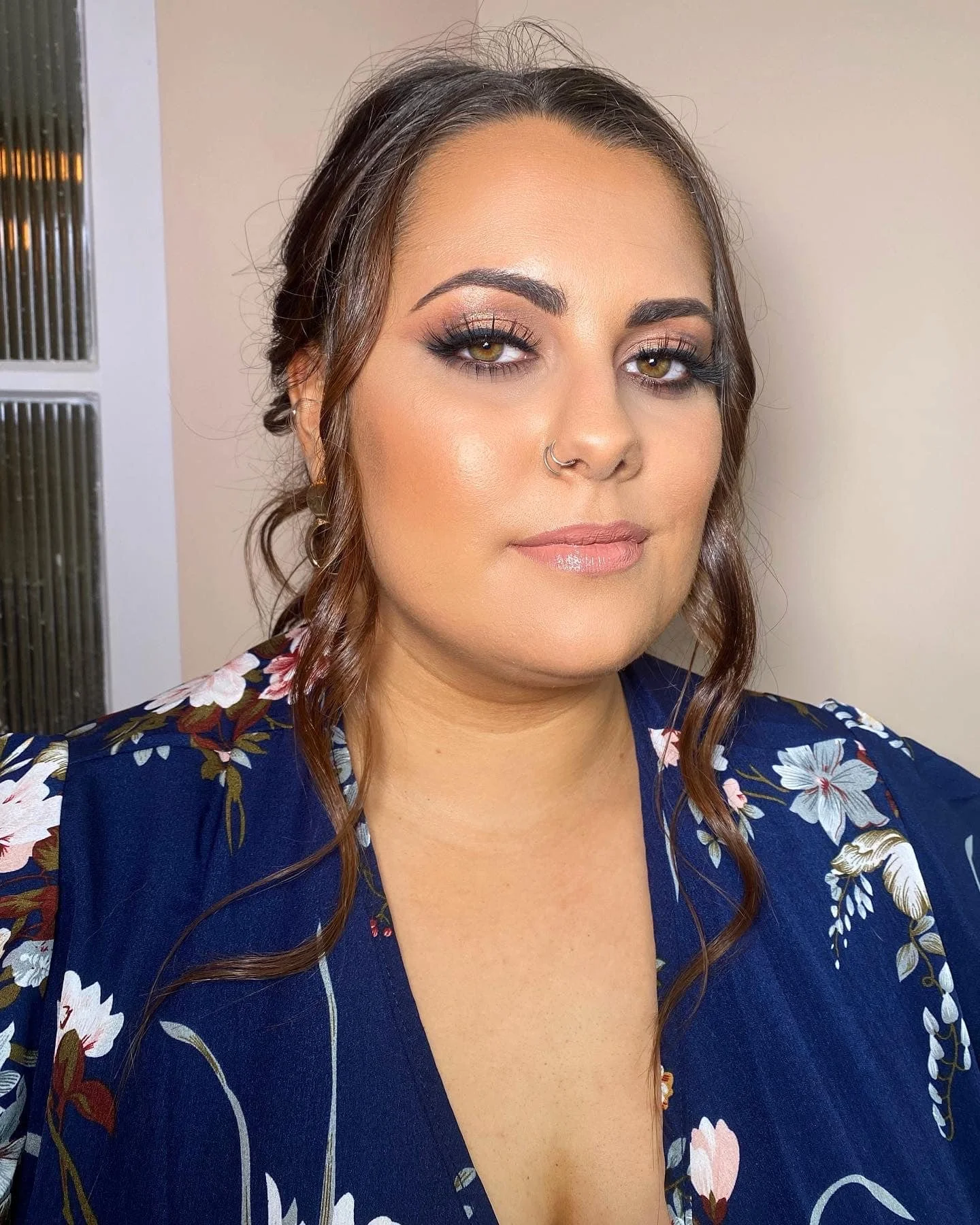 A woman with brown wavy hair, makeup, and a nose ring taking a selfie indoors. She is wearing a blue floral top.