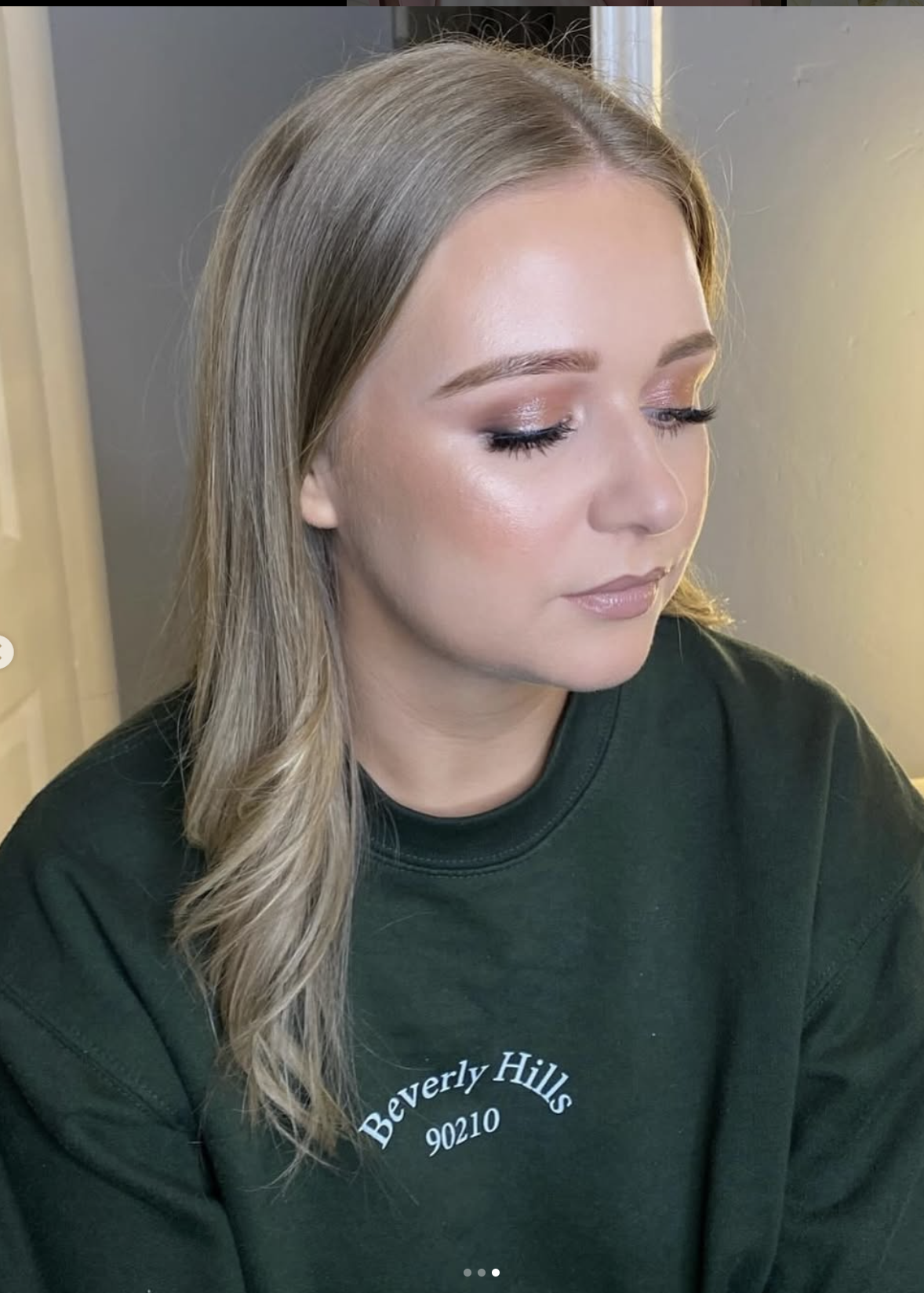 A woman with blonde hair, wearing a dark green Beverly Hills sweatshirt, looking down with closed eyes, makeup and natural lighting.