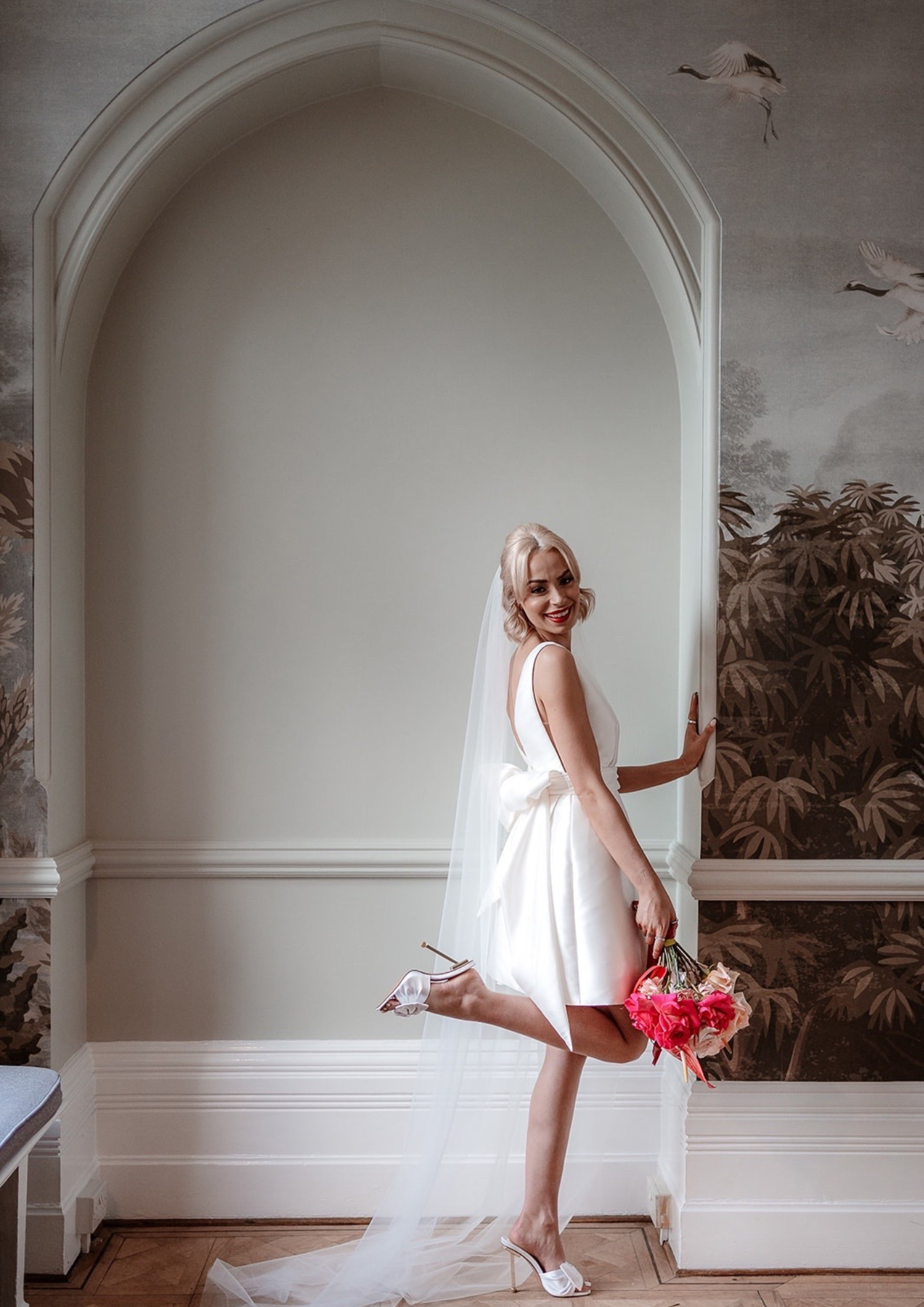 A smiling bride in a white wedding dress holding a bouquet of pink flowers, standing indoors against a decorative wall with an arch and botanical murals.