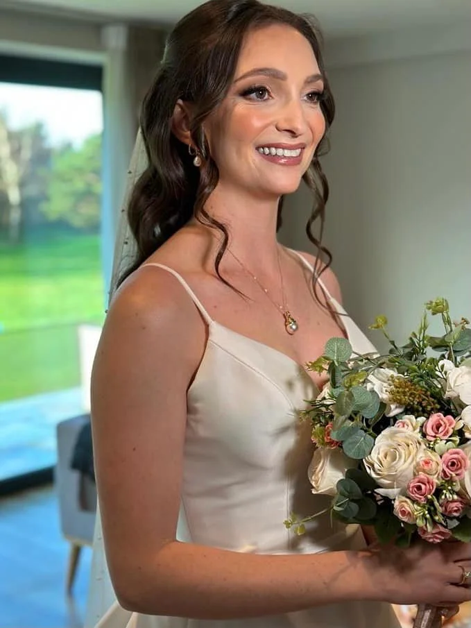 A woman with brown hair and light makeup in a wedding dress holding a bouquet of pink and white roses in a bright indoor setting.