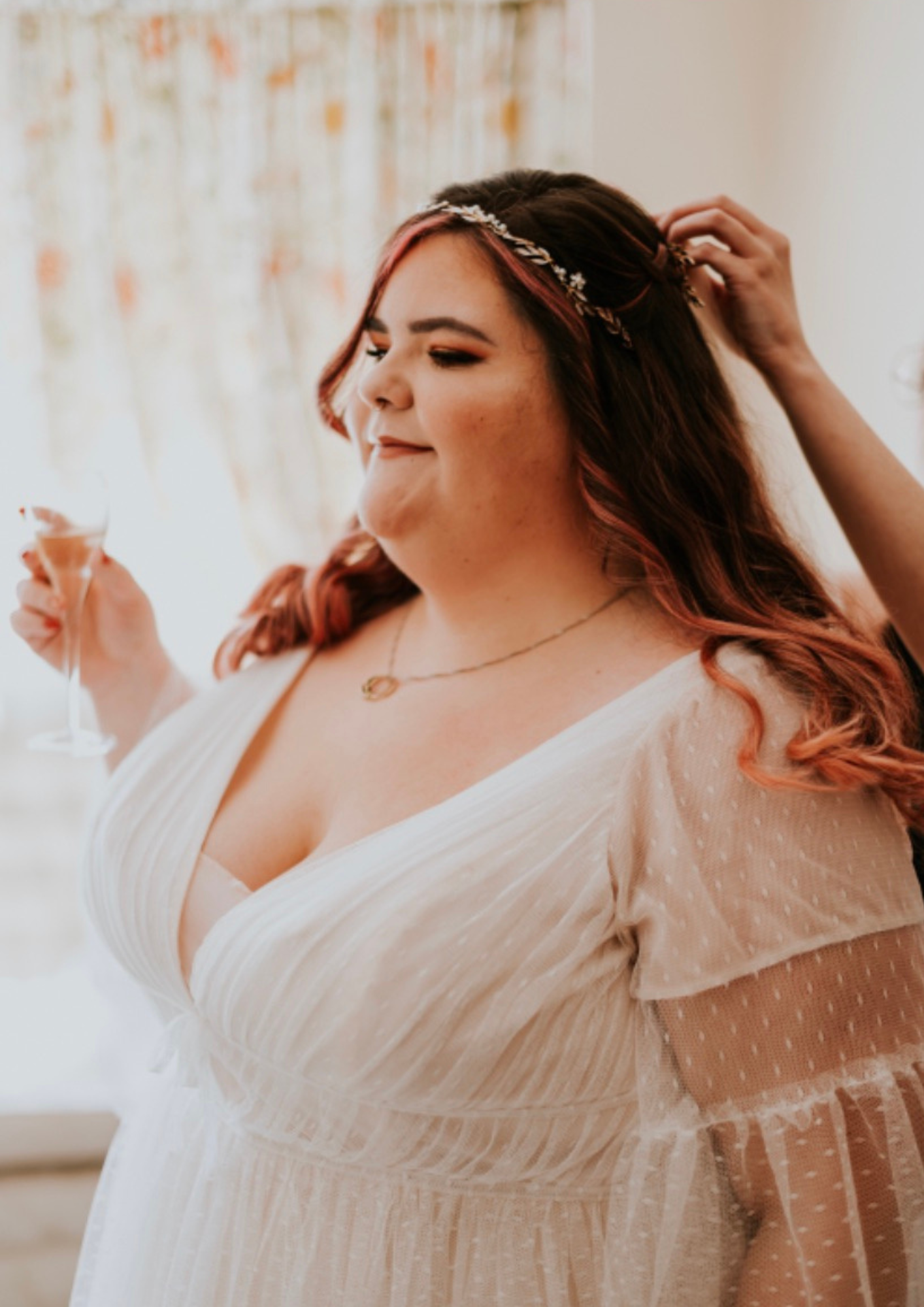 A woman with long, wavy hair with pink highlights, wearing a cream-colored, polka dot dress with sheer sleeves, is adjusting her hair while holding a glass of champagne, during a celebration or special occasion.