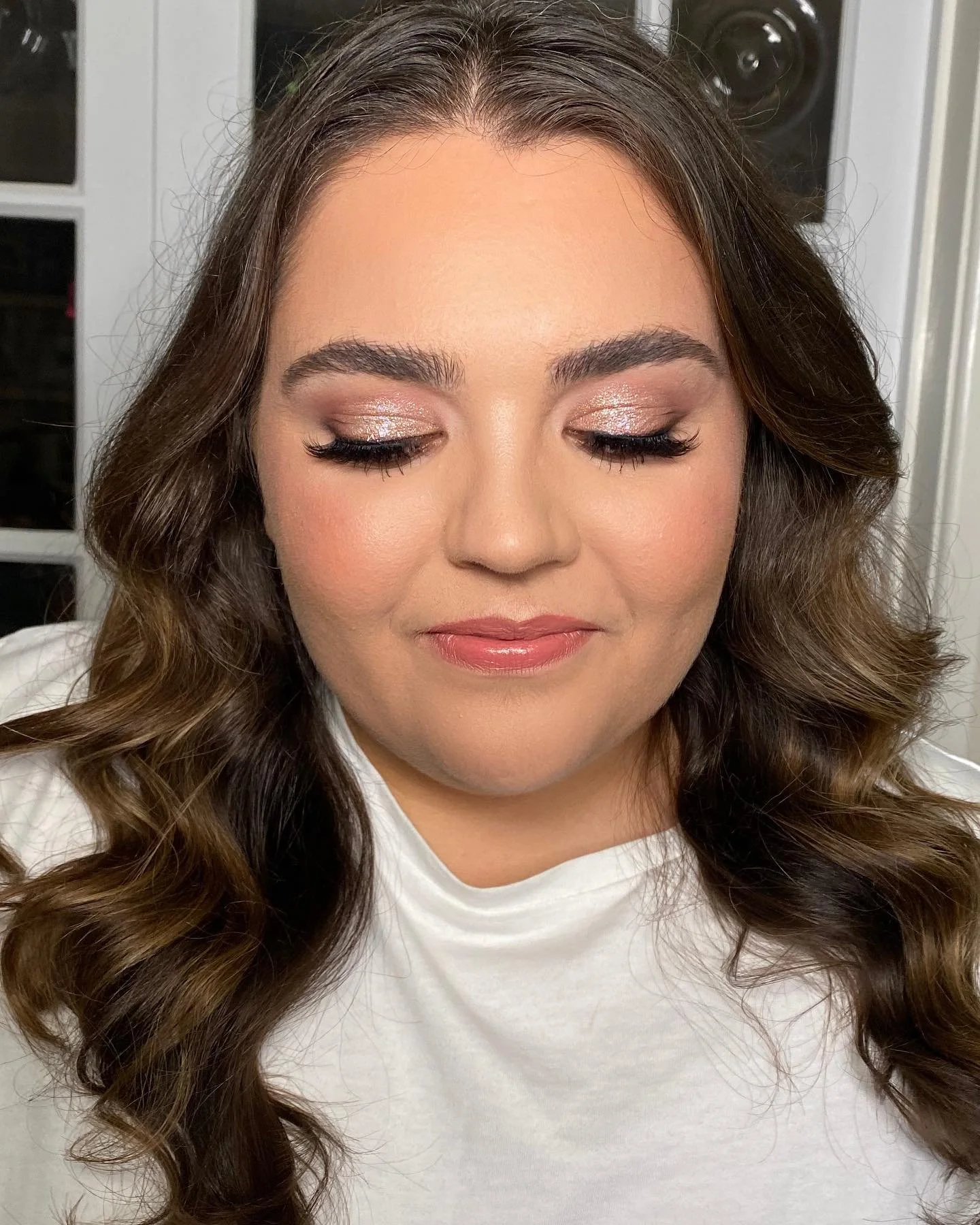 Close-up of a woman with medium brown, wavy hair and closed eyes showing shimmery eyeshadow and long eyelashes, wearing a white shirt, with a neutral expression.
