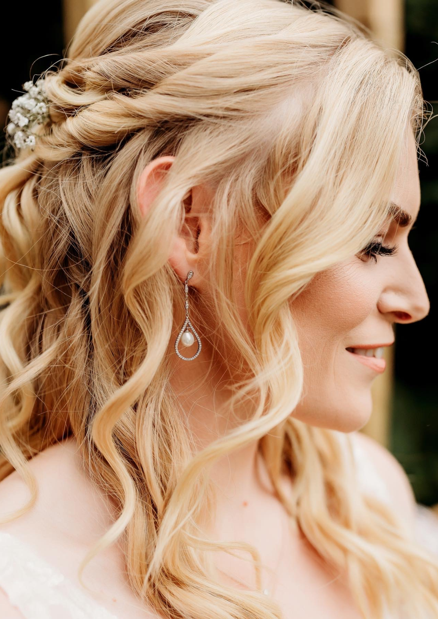 Close-up side profile of a blonde woman with wavy hair styled with baby’s breath flowers, wearing a pearl and diamond drop earring, smiling, and dressed in white.