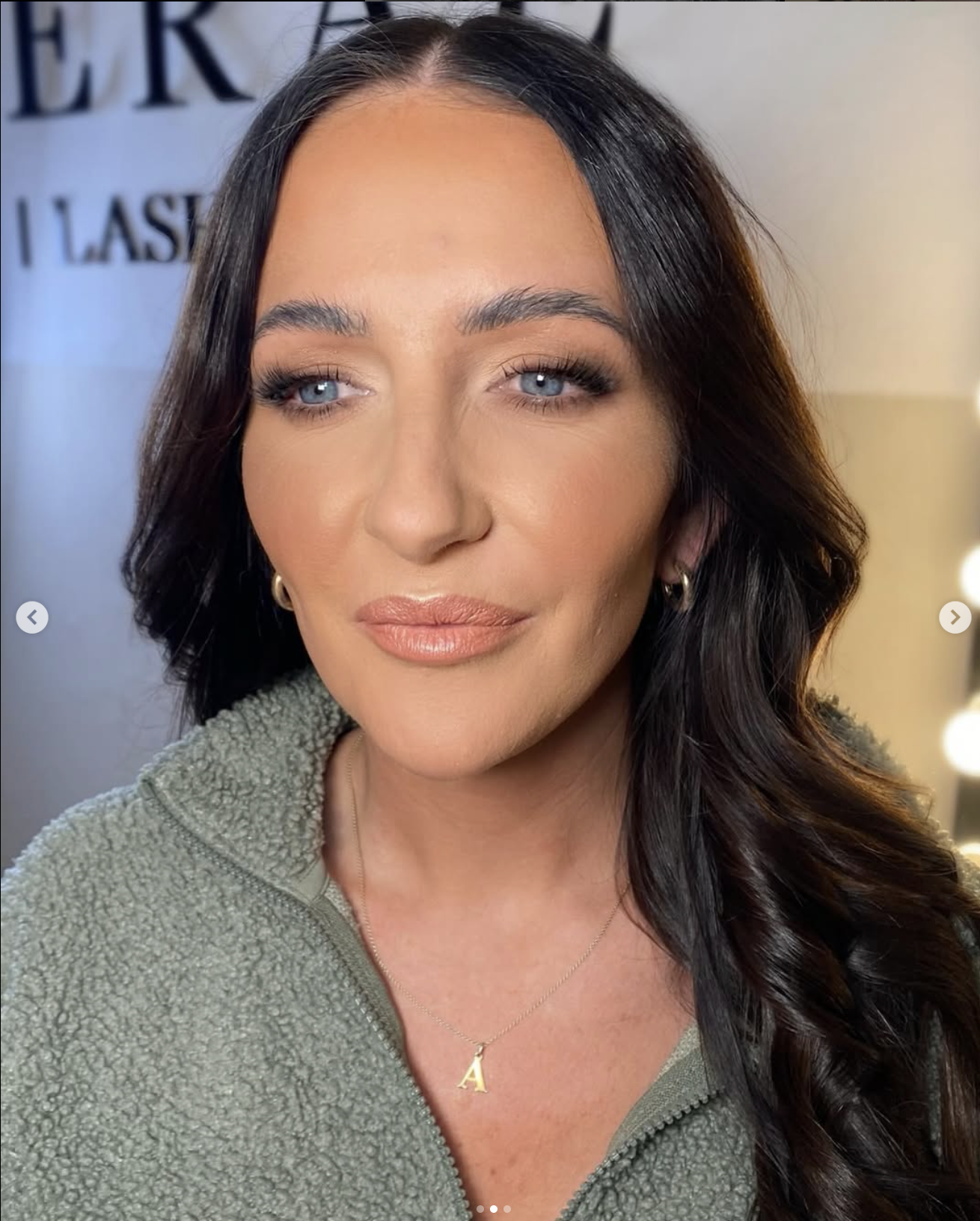 A woman with long wavy black hair, blue eyes, and makeup, wearing a green fuzzy zip-up jacket and a gold necklace with the letter A, smiling softly with a neutral background.