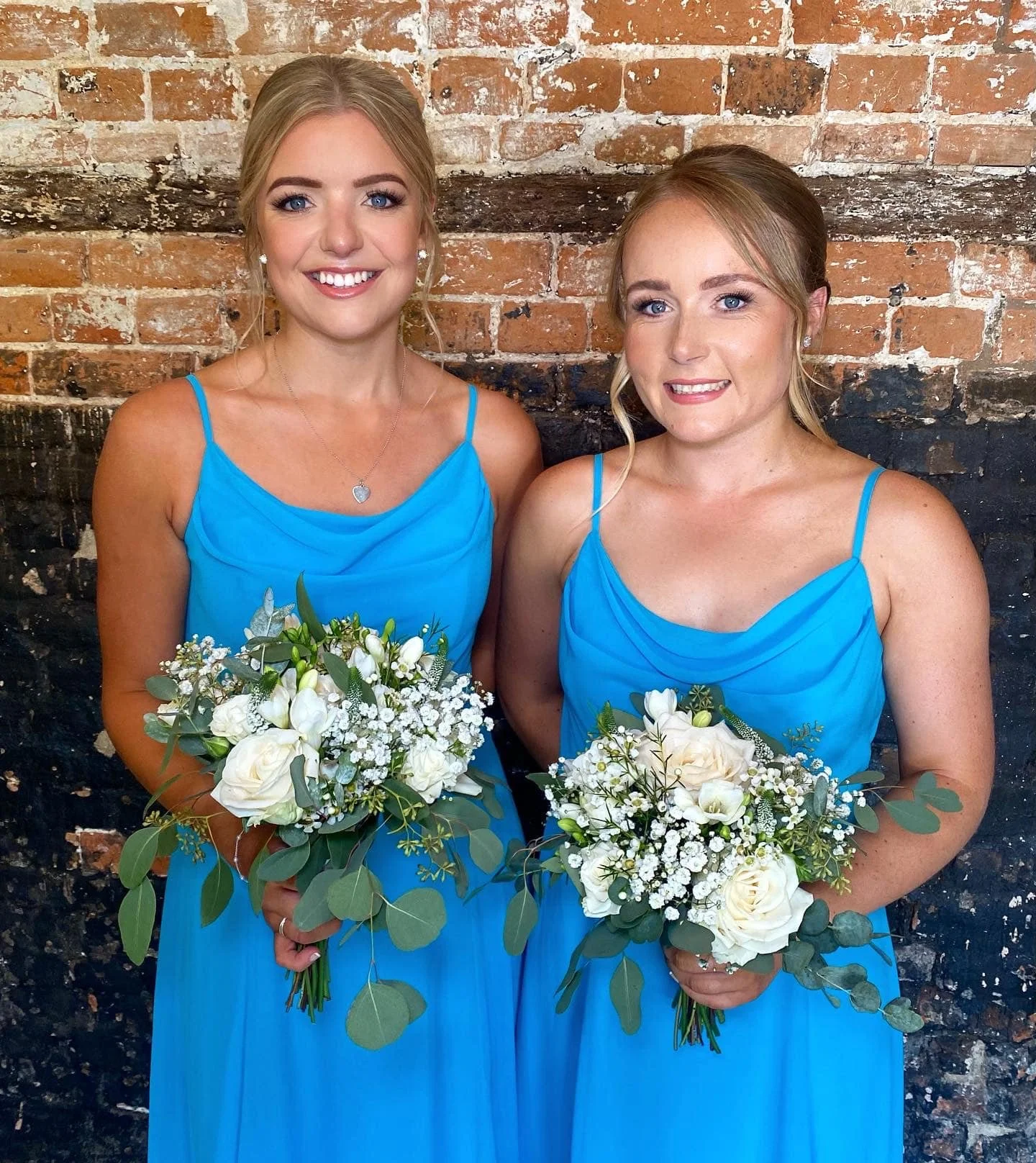 Two women in matching blue bridesmaid dresses holding bouquets of white flowers and greenery, standing against a brick wall.