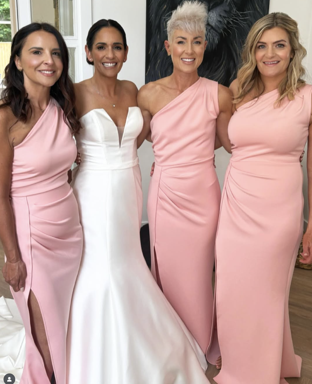 Four women dressed in pink bridesmaid dresses and one woman in a white wedding dress, smiling indoors with a black and white artwork behind them.
