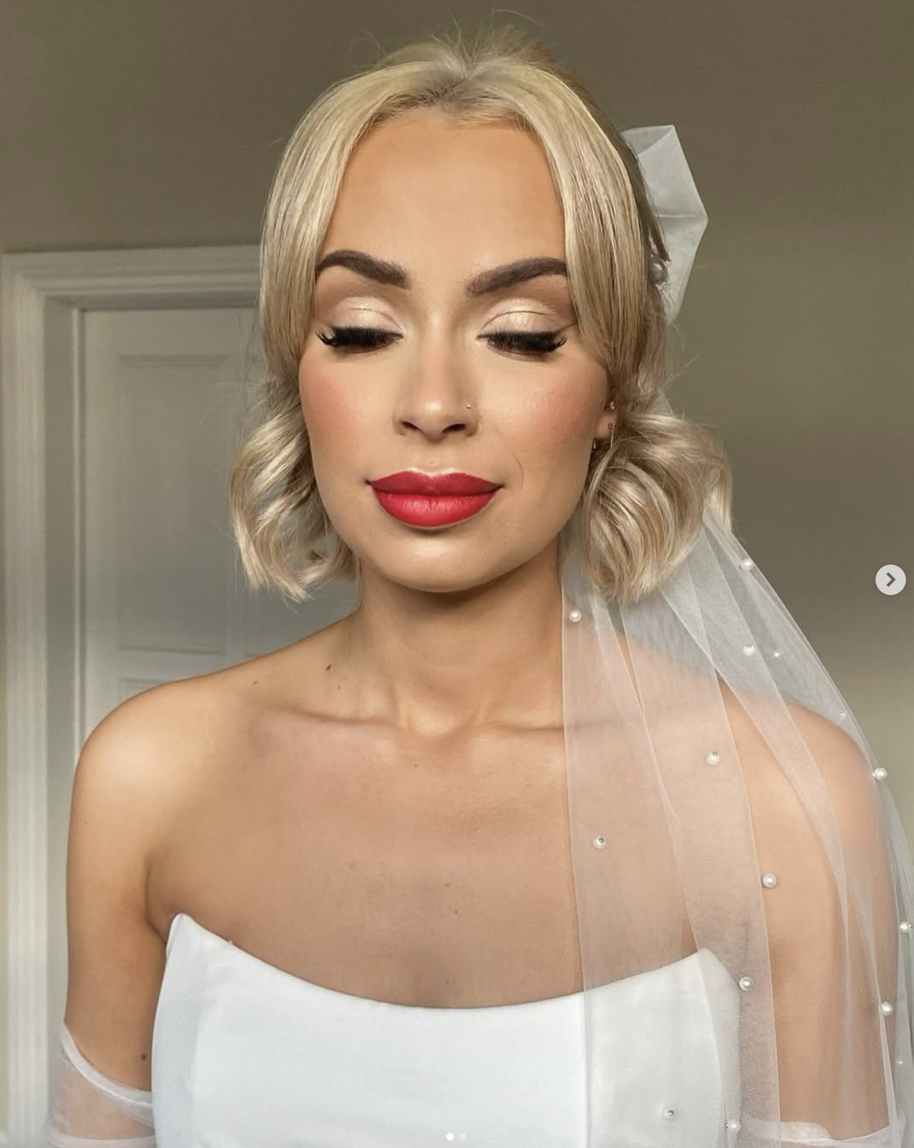 A woman with blonde, wavy, shoulder-length hair wearing a white dress and a sheer veil with pearls, with makeup including red lipstick, eyeshadow, and false eyelashes, standing indoors against a neutral background.