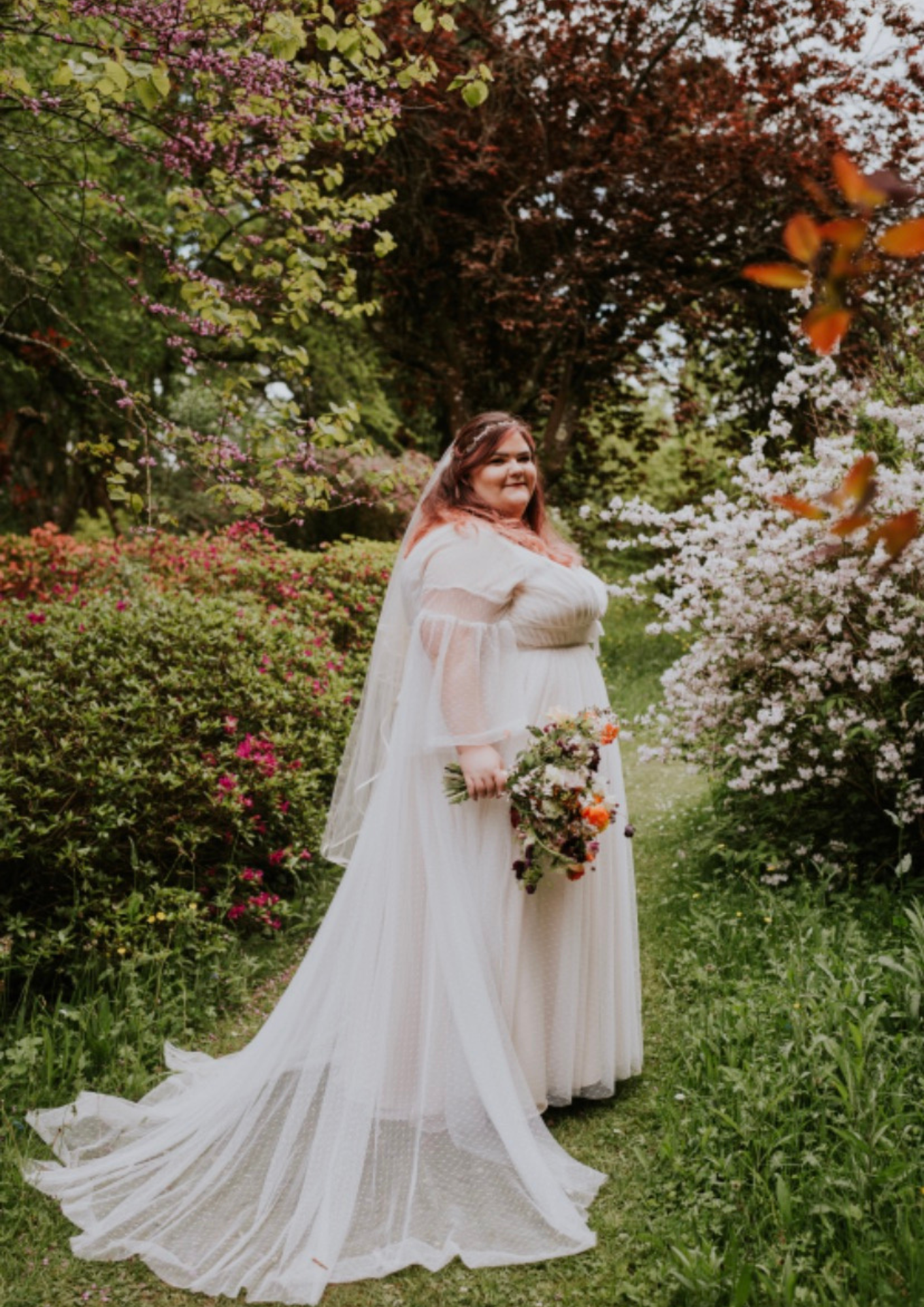 A woman in a white gown holding a wedding bouquet stands in a garden with blooming pink and white flowers and green trees.