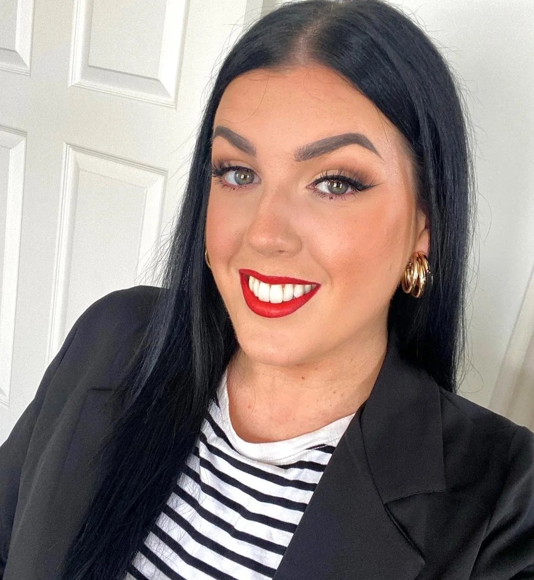 A woman with long black hair, wearing red lipstick, gold hoop earrings, a black blazer, and a black-and-white striped shirt, smiling in front of a white door.
