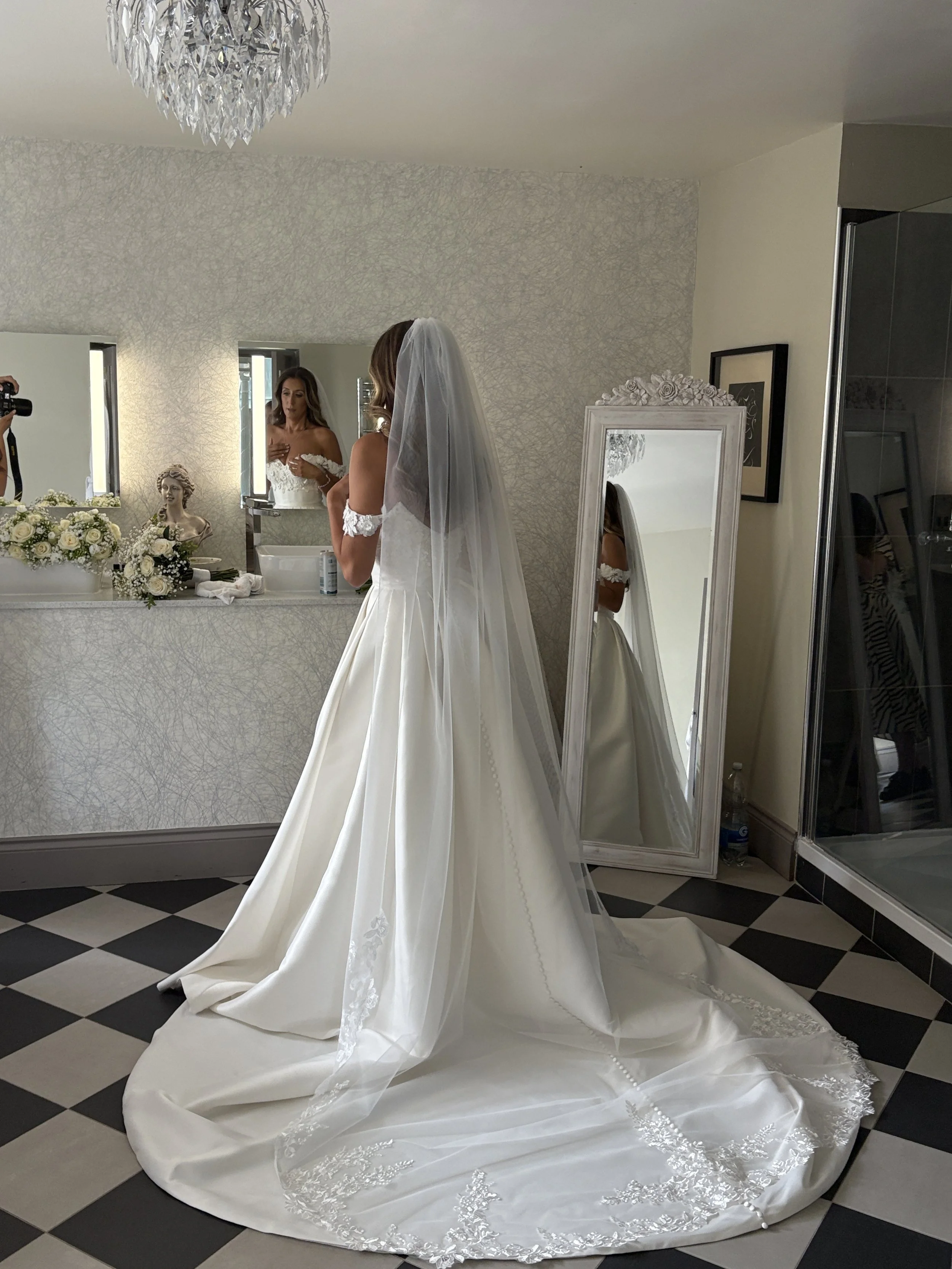 Bride in a white wedding dress with a long train and veil, looking in a mirror while getting ready in a room decorated with floral arrangements and a chandelier.