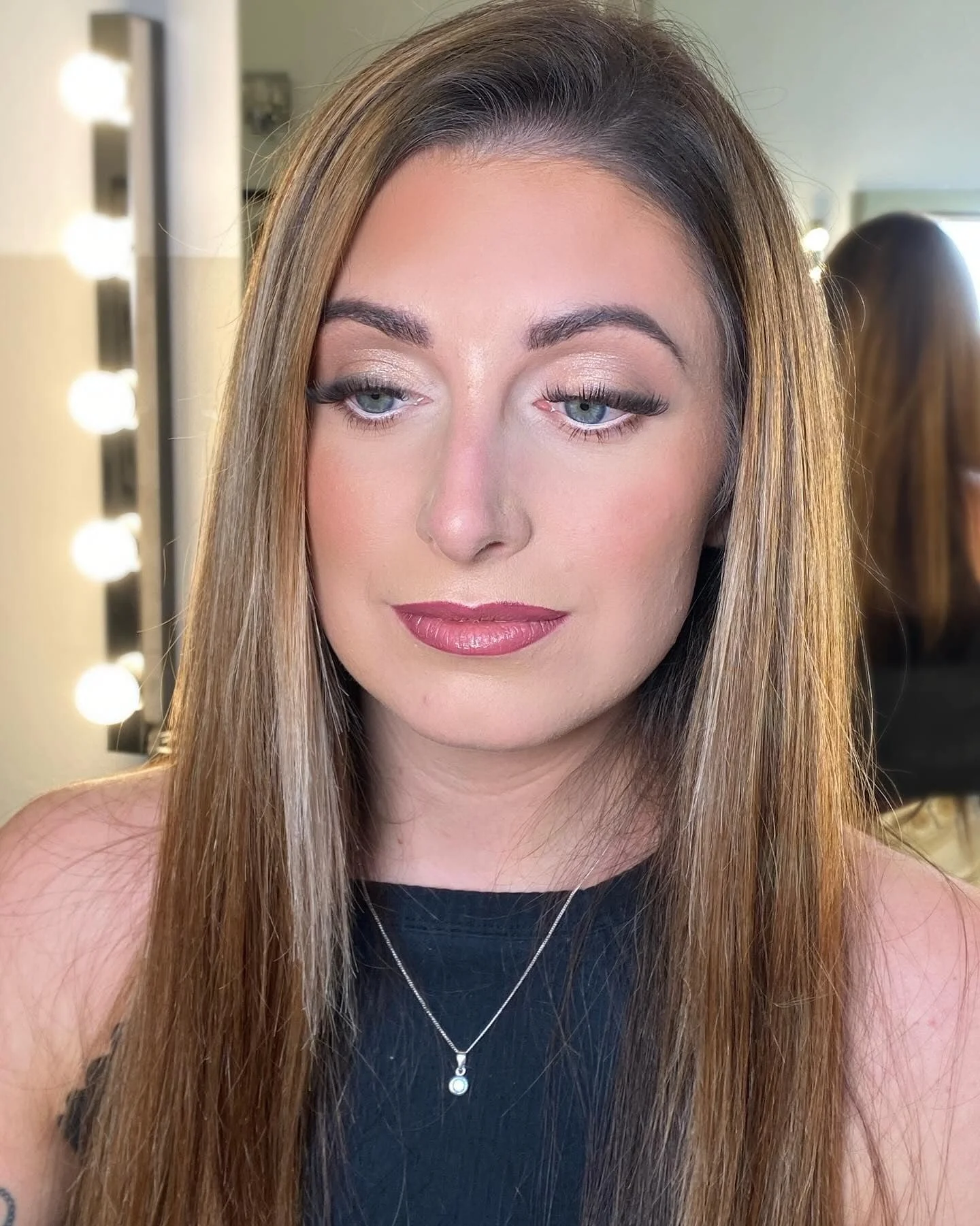 Close-up portrait of a woman with long brown hair, makeup, and a necklace, in a dressing room with bright vanity lights.